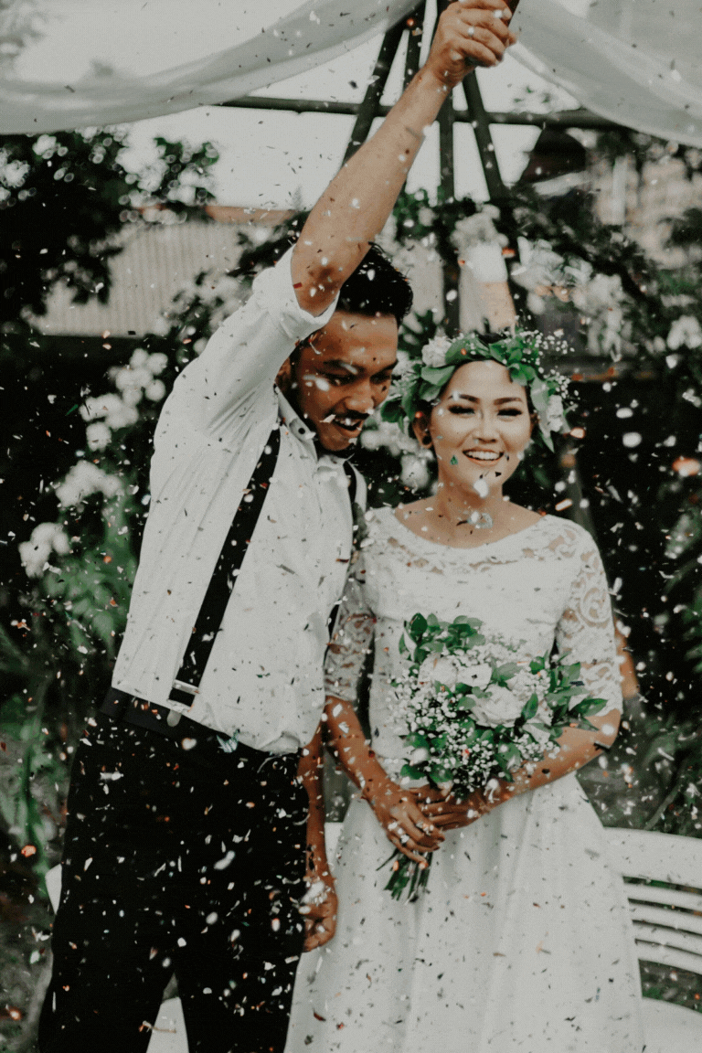 A newly married couple celebrating outdoors with confetti, the groom lifting his arm and the bride holding a bouquet, both smiling.