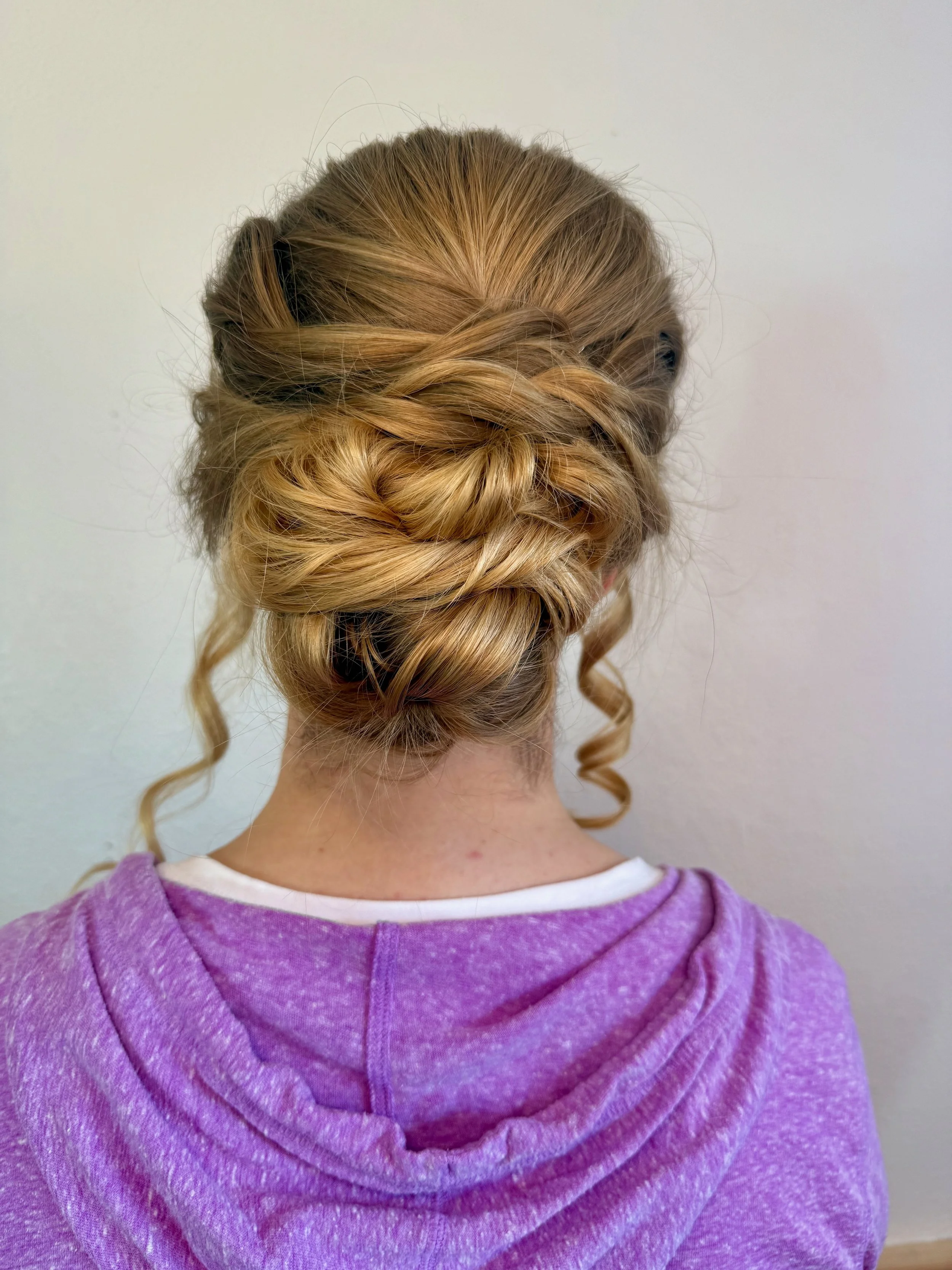 Back of a bride with blonde hair styled in an elegant updo with loose curls, wearing a purple hoodie.