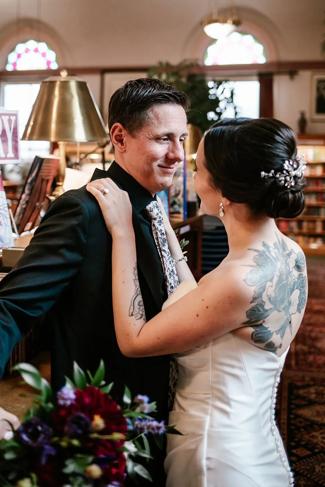 A bride and groom sharing a dance, with the bride wearing a white wedding gown and the groom in a black suit, in a warmly lit indoor setting with bookshelves and stained glass windows in the background.
