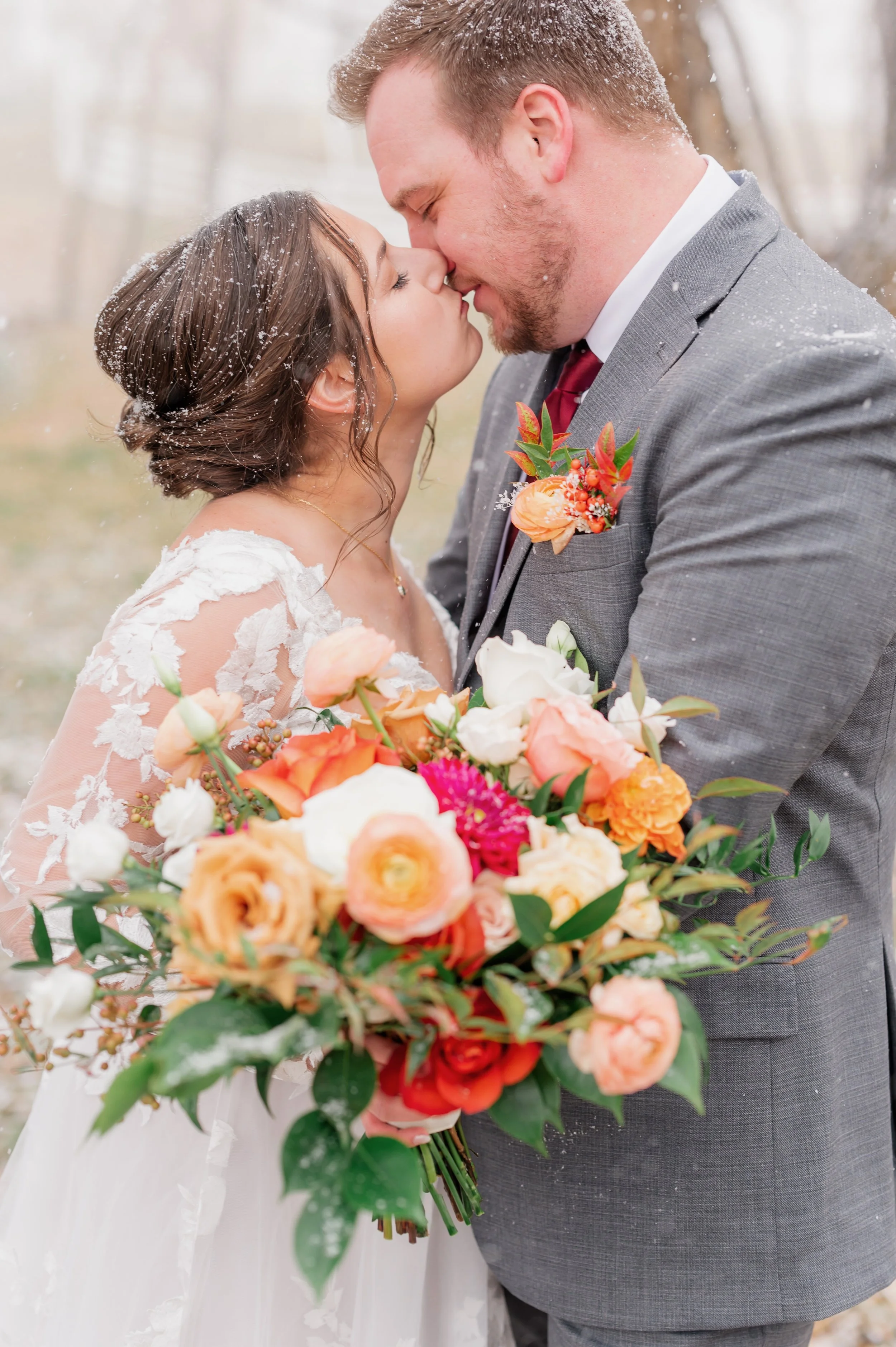 A bride and groom standing close, about to kiss, outside in a snowy setting, holding a large colorful bouquet of flowers.