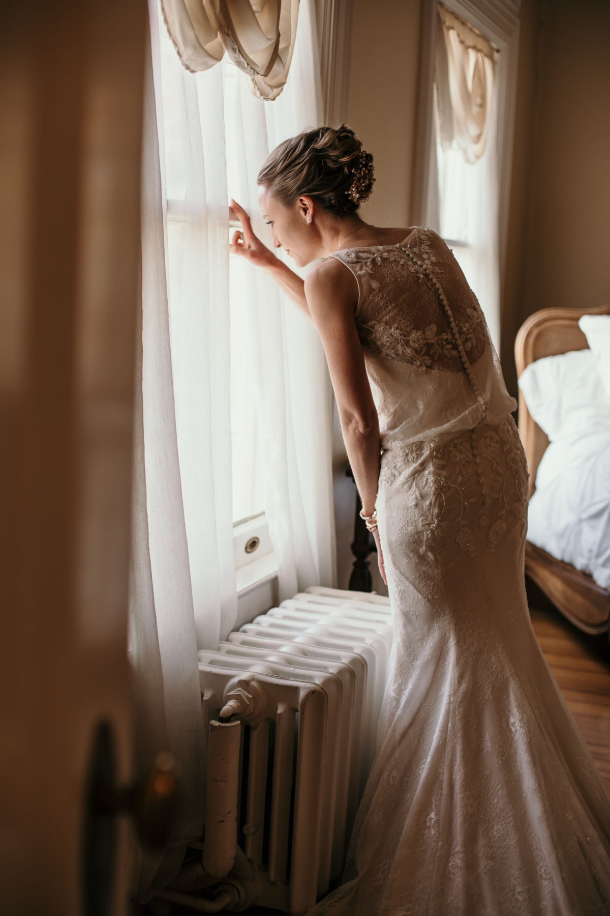 A bride in a lace wedding gown standing by a window, looking out and touching the curtain.