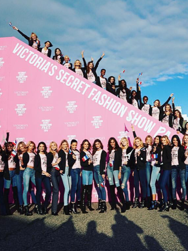 Group of women preparing to board a plane for the Paris Victoria's Secret Fashion Show, some standing in front and others sitting or standing on the pink backdrop, waving flags and smiling.