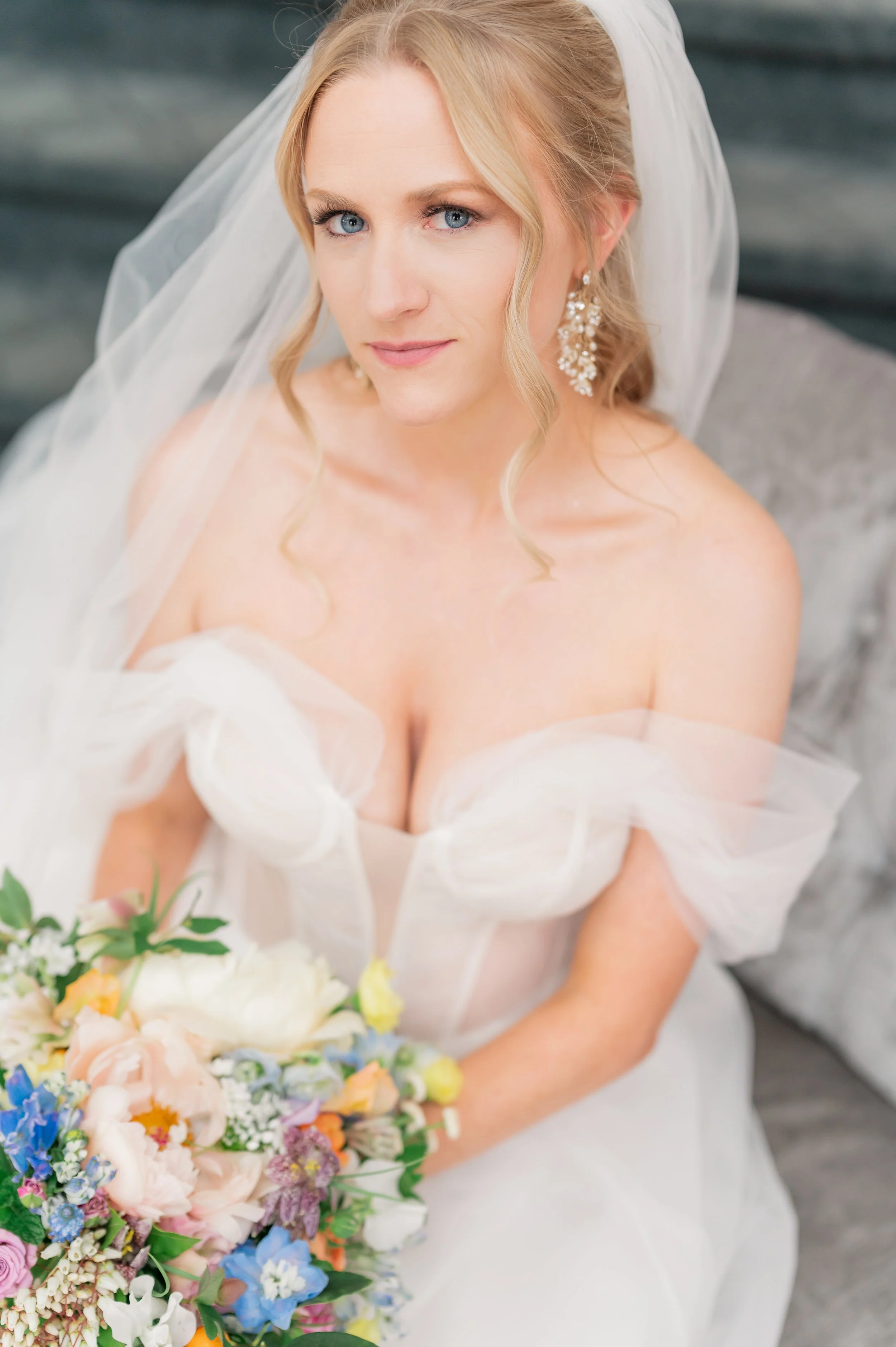 A bride with blonde hair and blue eyes, wearing a white strapless wedding dress and large pearl earrings, holding a colorful bouquet of flowers.