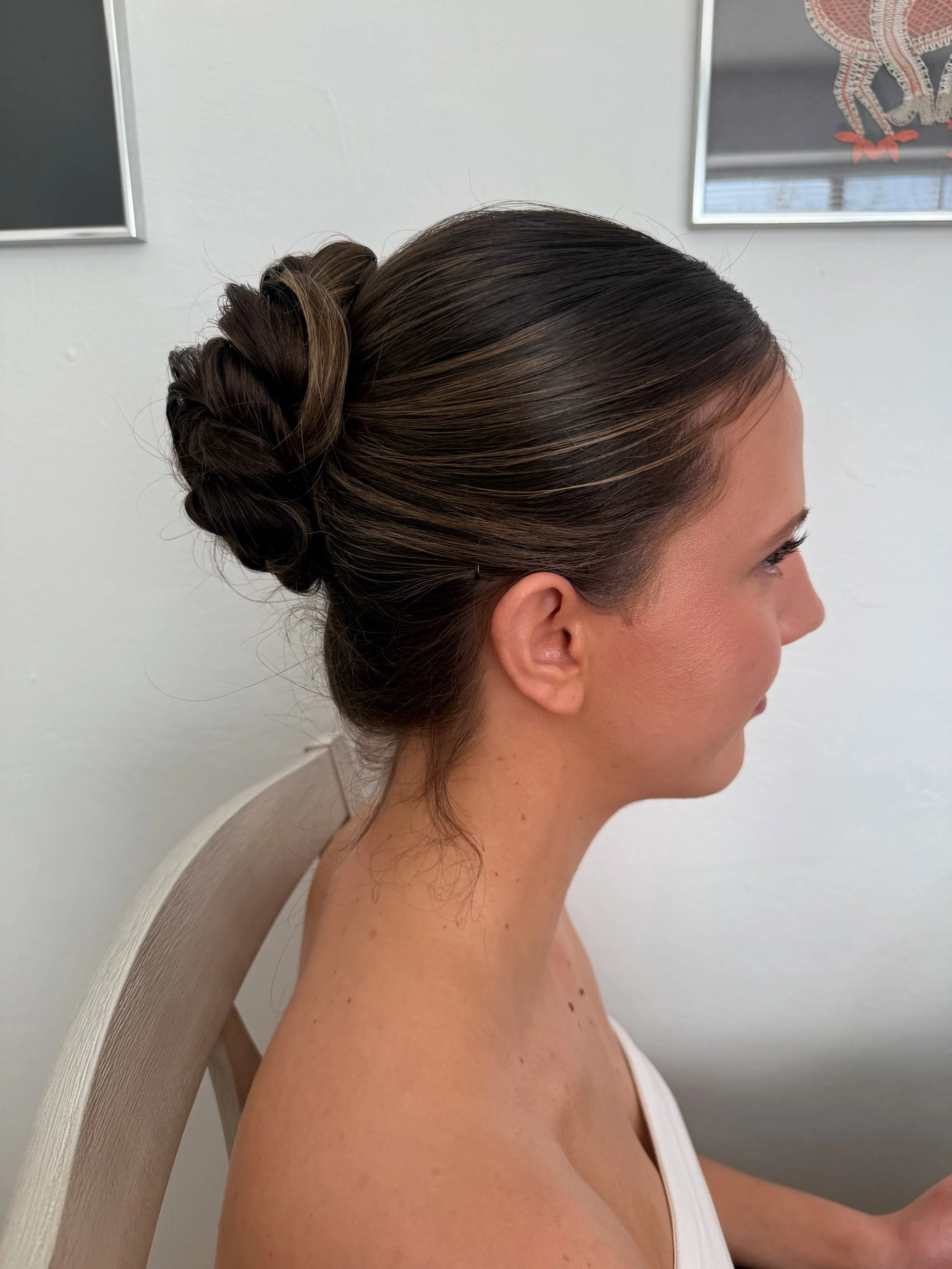 A bride with dark, styled hair sitting on a beige chair against a white wall, with a framed art piece and a window in the background.