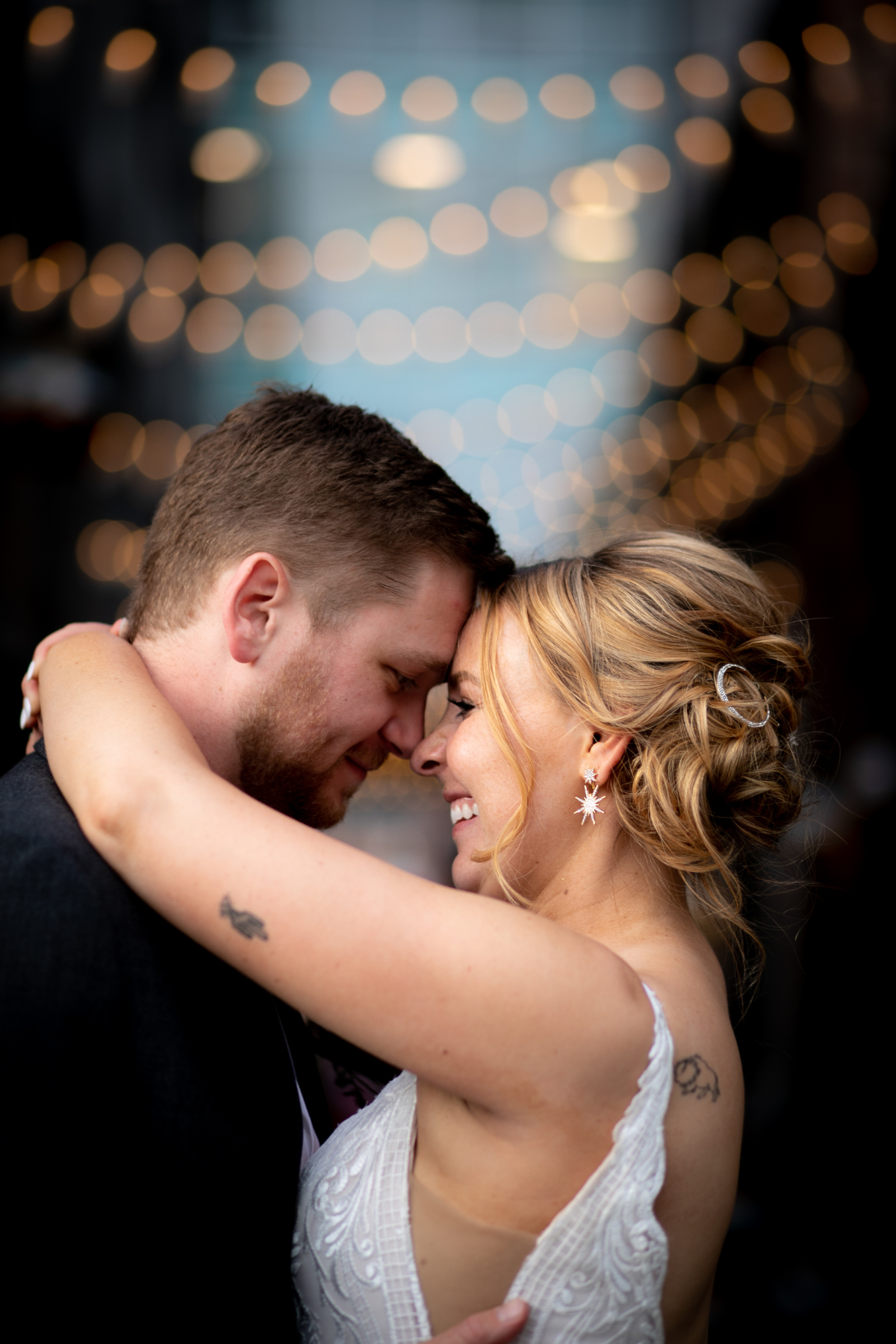 A couple sharing an intimate moment, with their foreheads and noses touching, embracing each other and smiling, at a celebration or wedding with blurred string lights in the background.