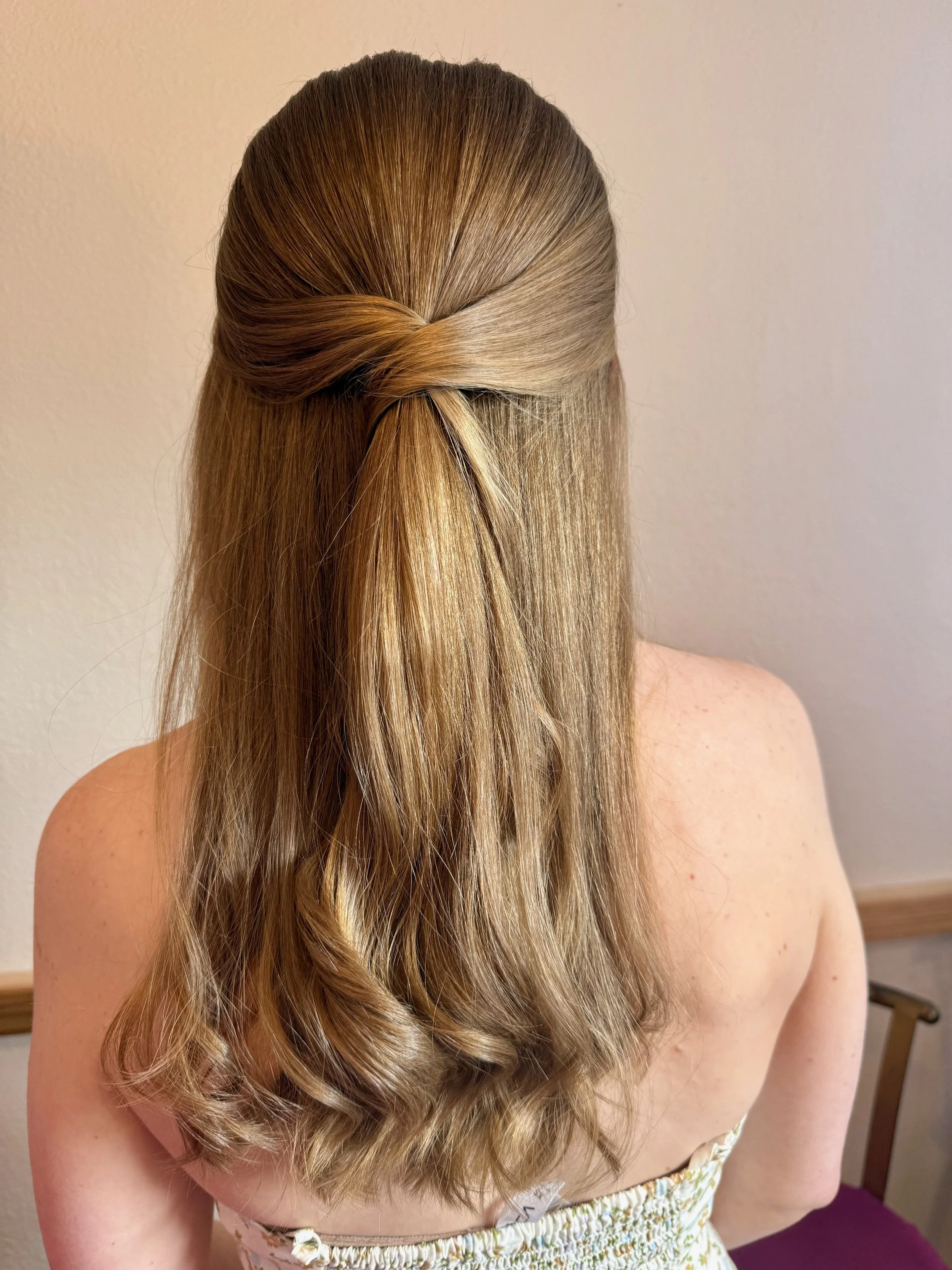 Back view of a bride with long, wavy, blonde hair styled in a half-updo with a twist, standing indoors against a plain wall.