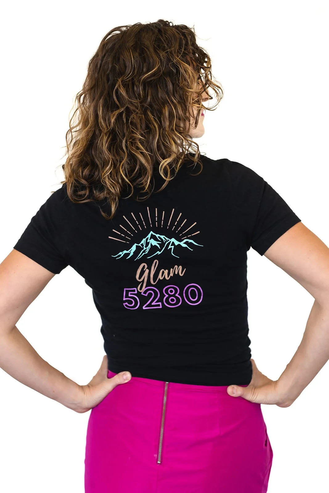 Back view of a woman with curly hair wearing a black t-shirt with a mountain graphic and the words 'Glam 5280' on the back, and a pink skirt with a visible zipper.