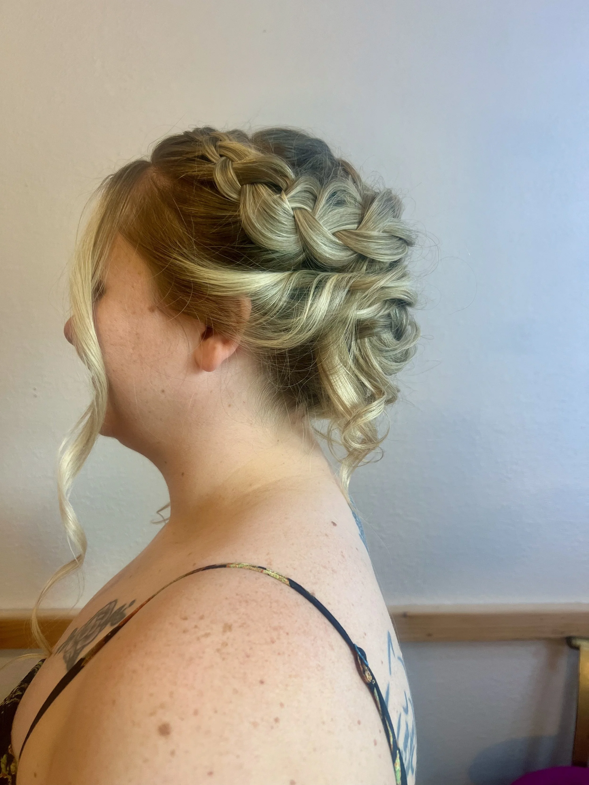 A bride's side profile with blonde hair styled in loose curls and a braided crown. She has freckles and is wearing a black spaghetti strap top, with a tattoo visible on her shoulder.