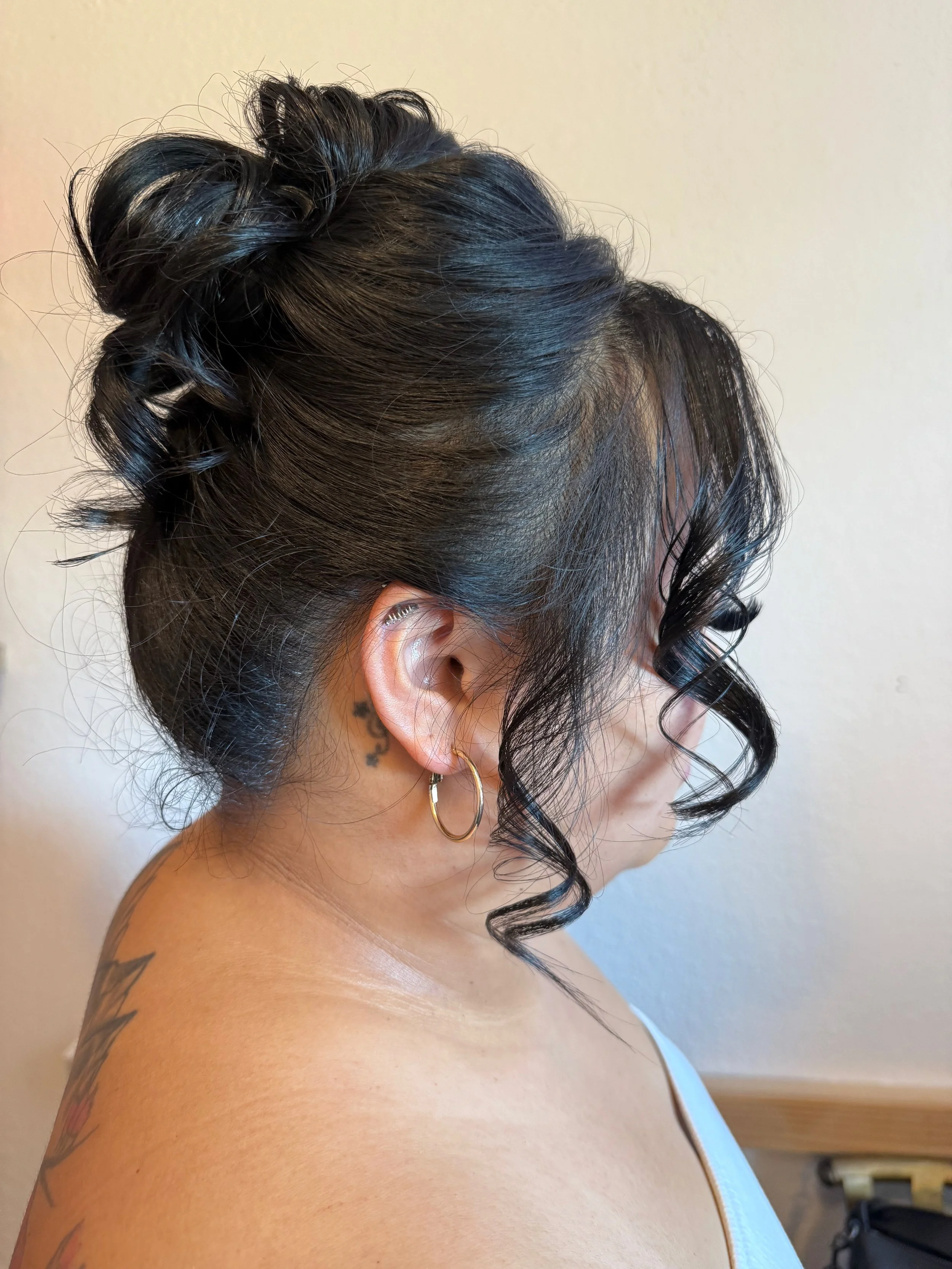 A bride with black curly hair styled in an updo with loose curls framing her face, wearing hoop earrings, and a tattoo on her shoulder.