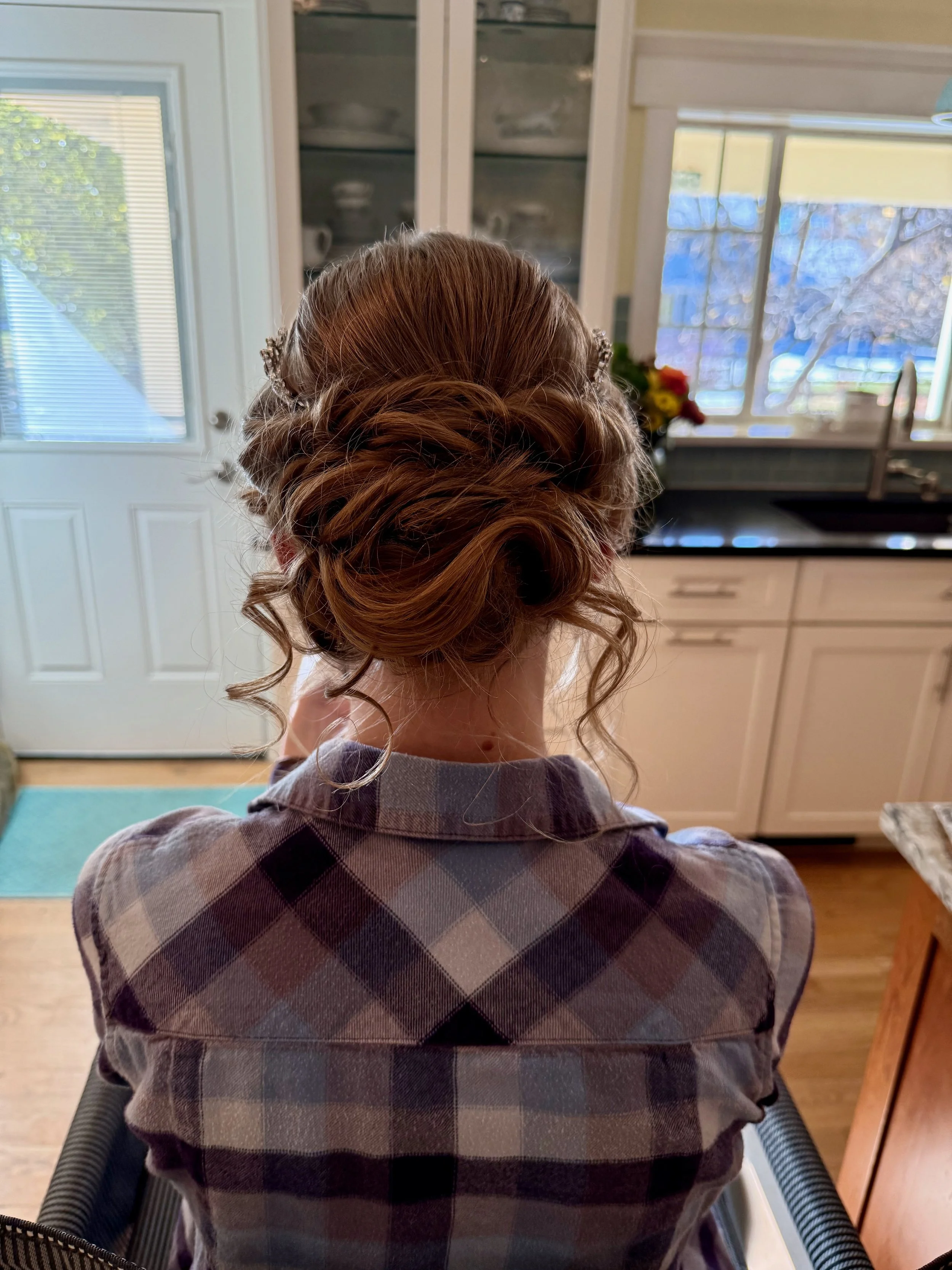 Back view of a bride with styled red hair in an updo, sitting in a kitchen, wearing a purple and black plaid shirt, with sunlight shining through a window and a door with blinds in the background.