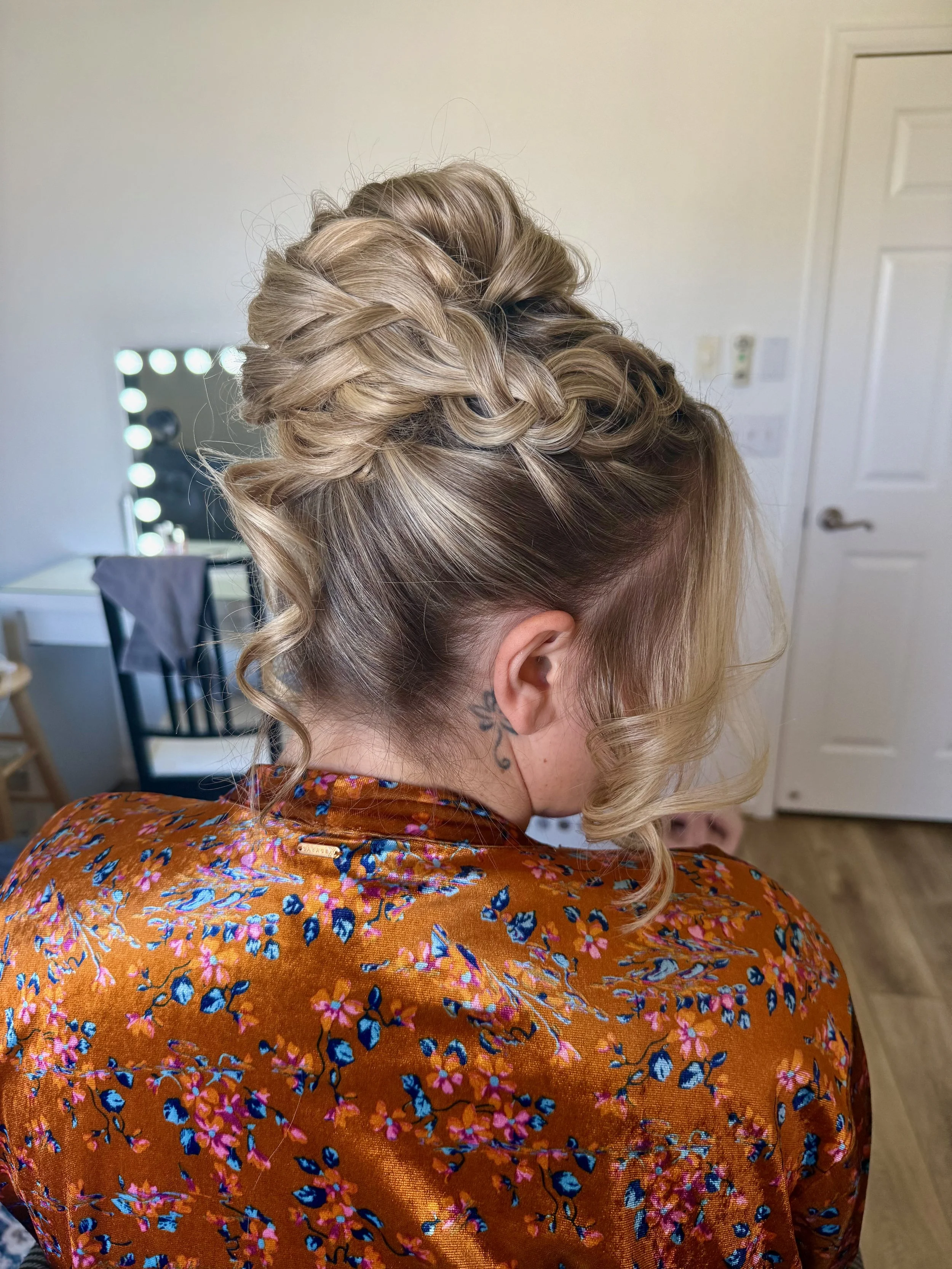 A bride with blonde hair styled in an elegant updo with braids and curls, wearing a floral orange dress, standing in a room with a mirror and makeup lights in the background.