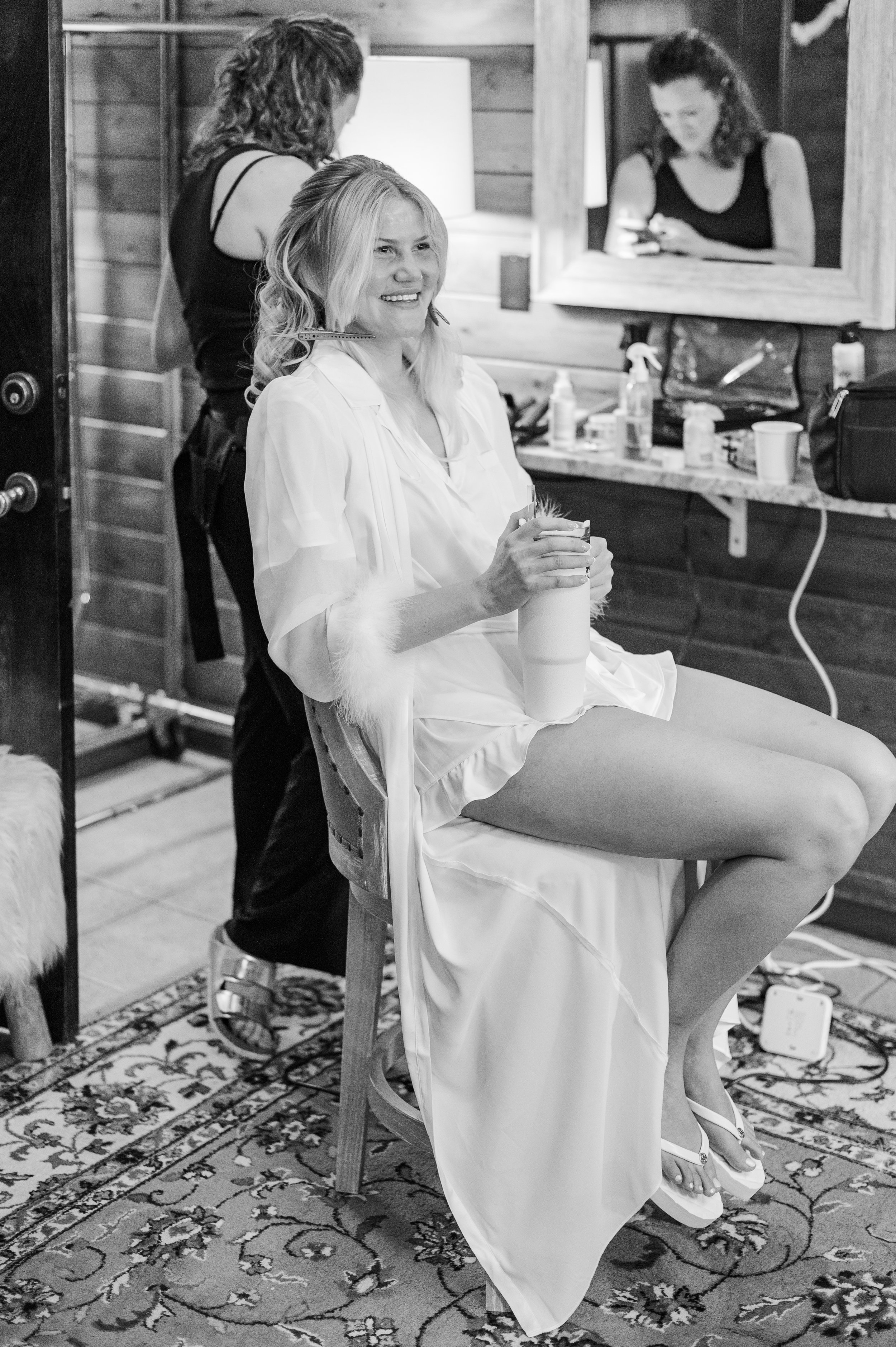 Woman in a white dress sitting on a chair with her legs crossed, smiling while having her hair styled by a hairstylist in a salon, with a mirror and various hair and cosmetic products on the counter behind her.
