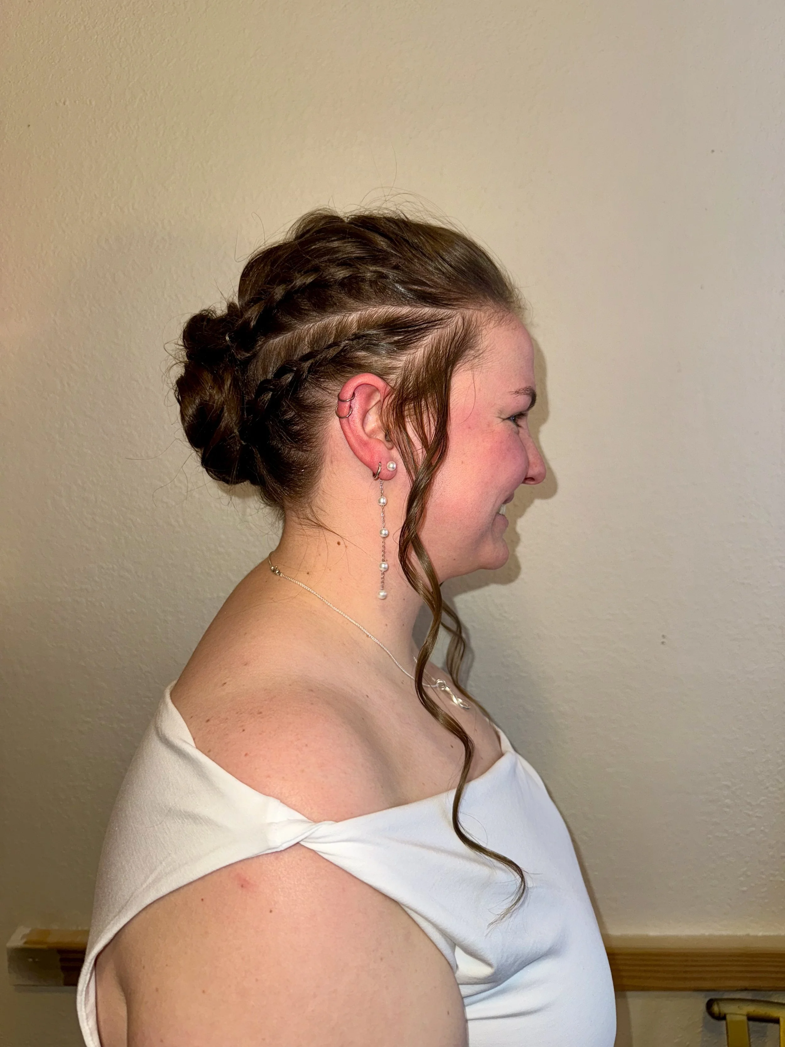 Side profile of a bride with viking braided hair, earrings, and a white top, standing against a beige wall.