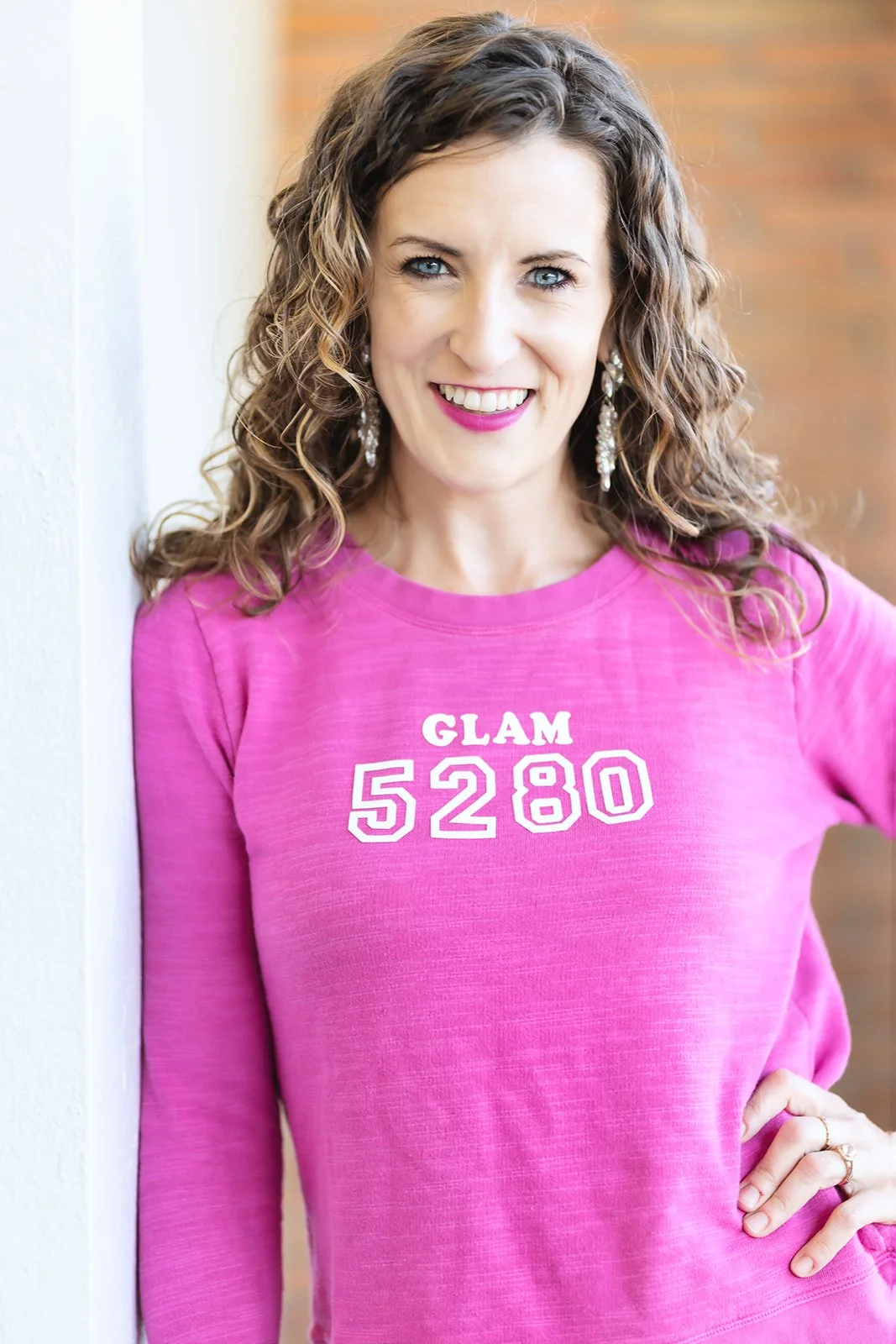A woman with curly brown hair and blue eyes smiling, wearing a pink sweatshirt with the words 'GLAM 5280' written on it, standing against a white wall with a brick background.