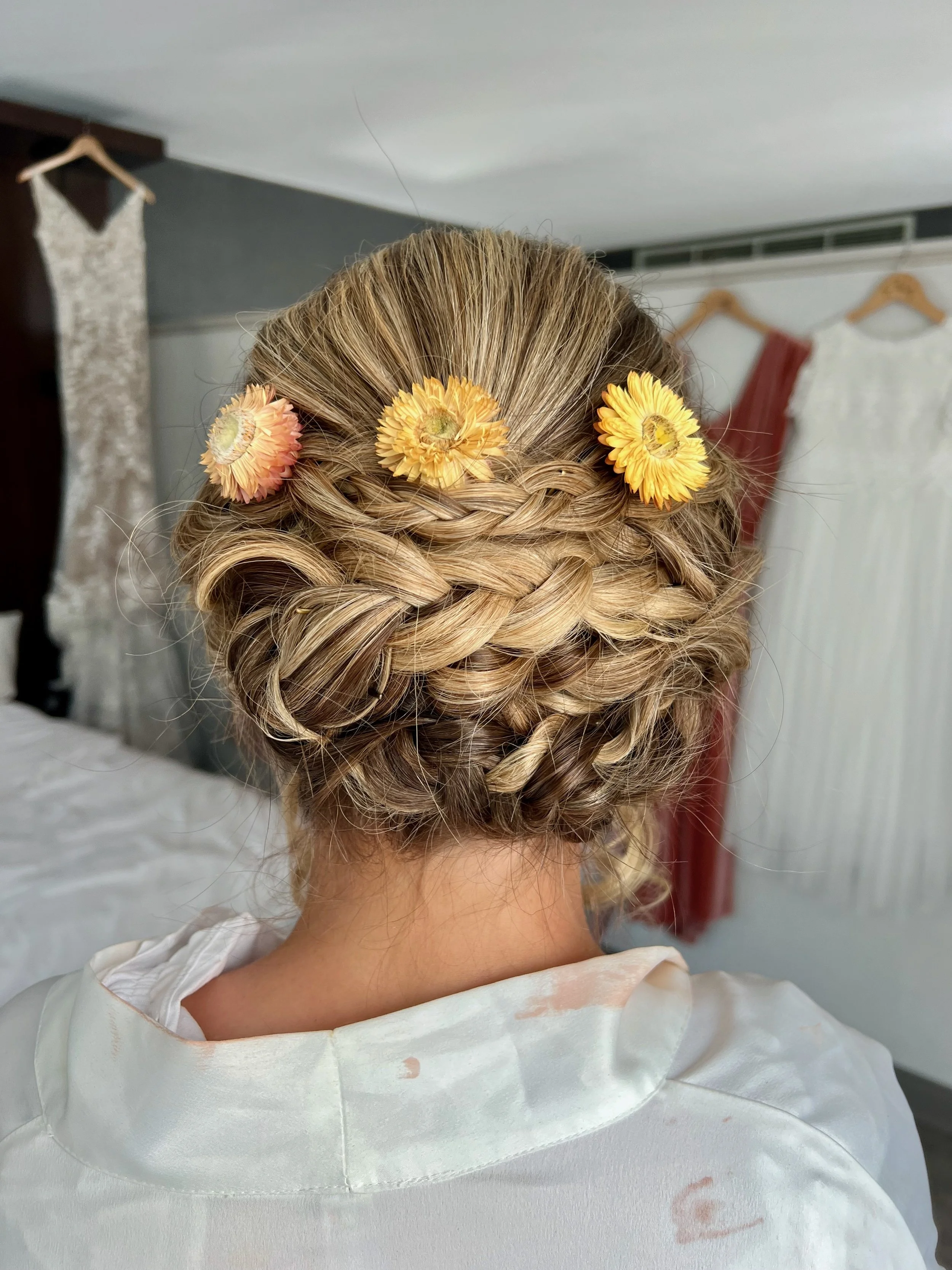 A bride with blonde hair styled in an intricate updo with braided sections, decorated with three yellow flowers, viewed from the back.