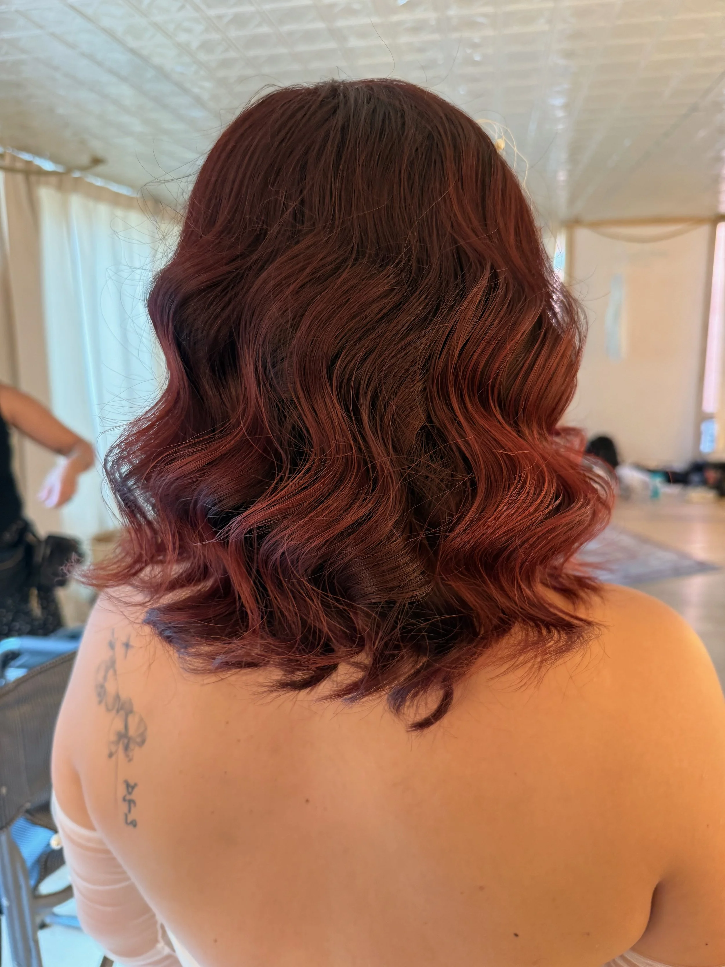 Back view of a bride with Hollywood wave, reddish-brown hair, partially visible tattoo on her upper arm, standing in a room with curtains and other people in the background.