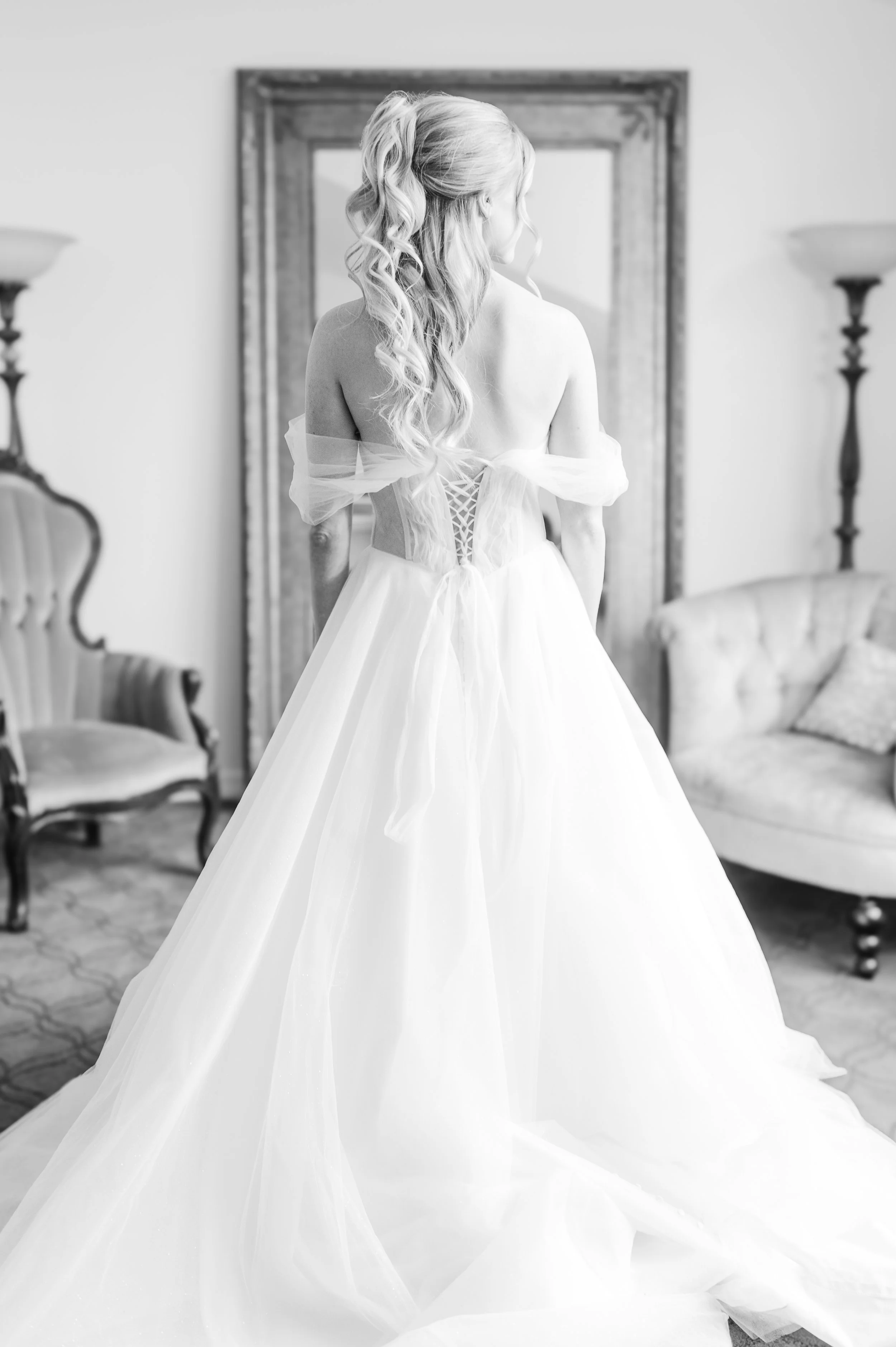 A woman in a wedding dress with long, curly hair, standing indoors and looking away from the camera.