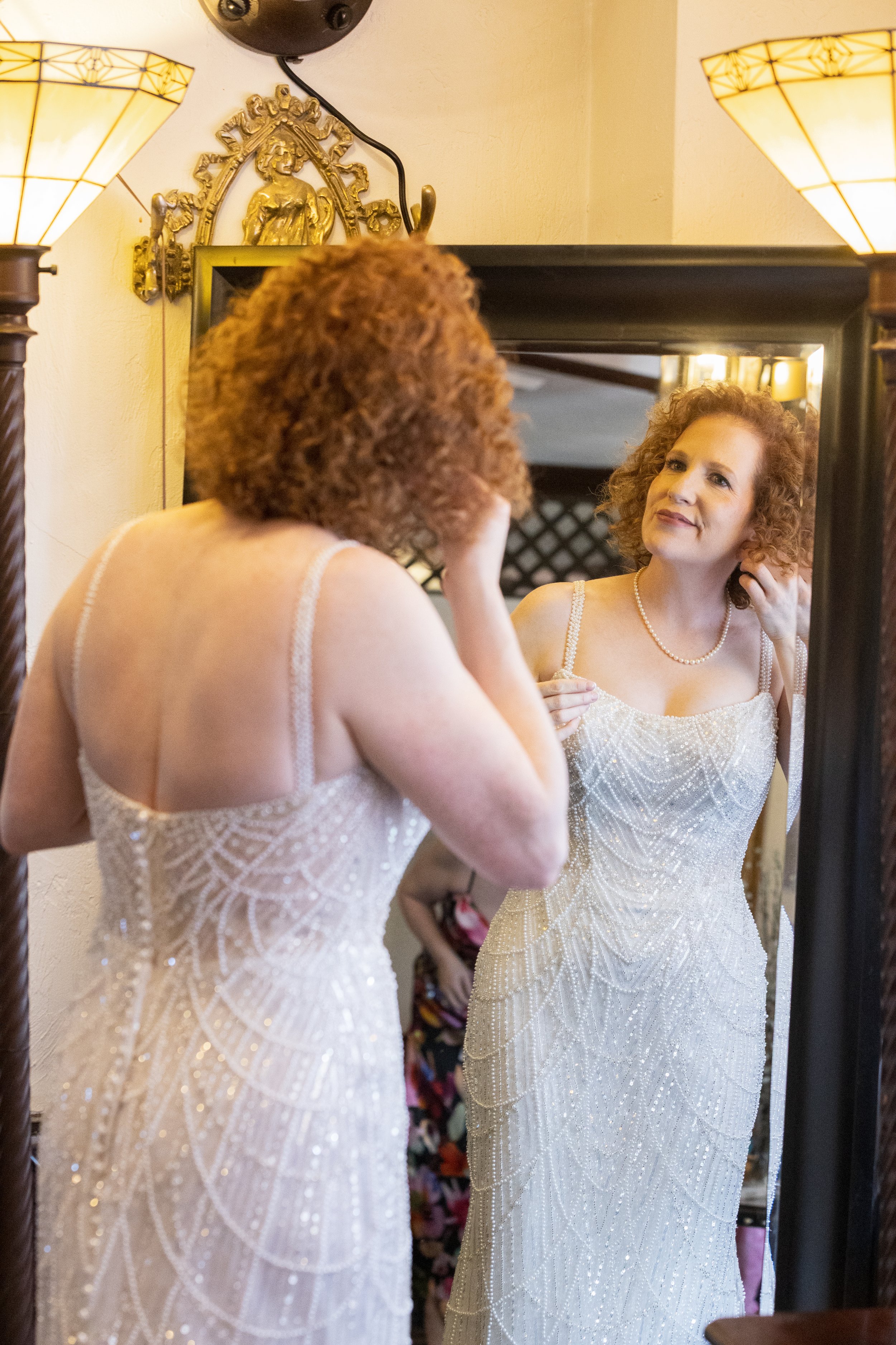 A bride with curly red hair looking at her reflection in a mirror, wearing a white, beaded dress with spaghetti straps.
