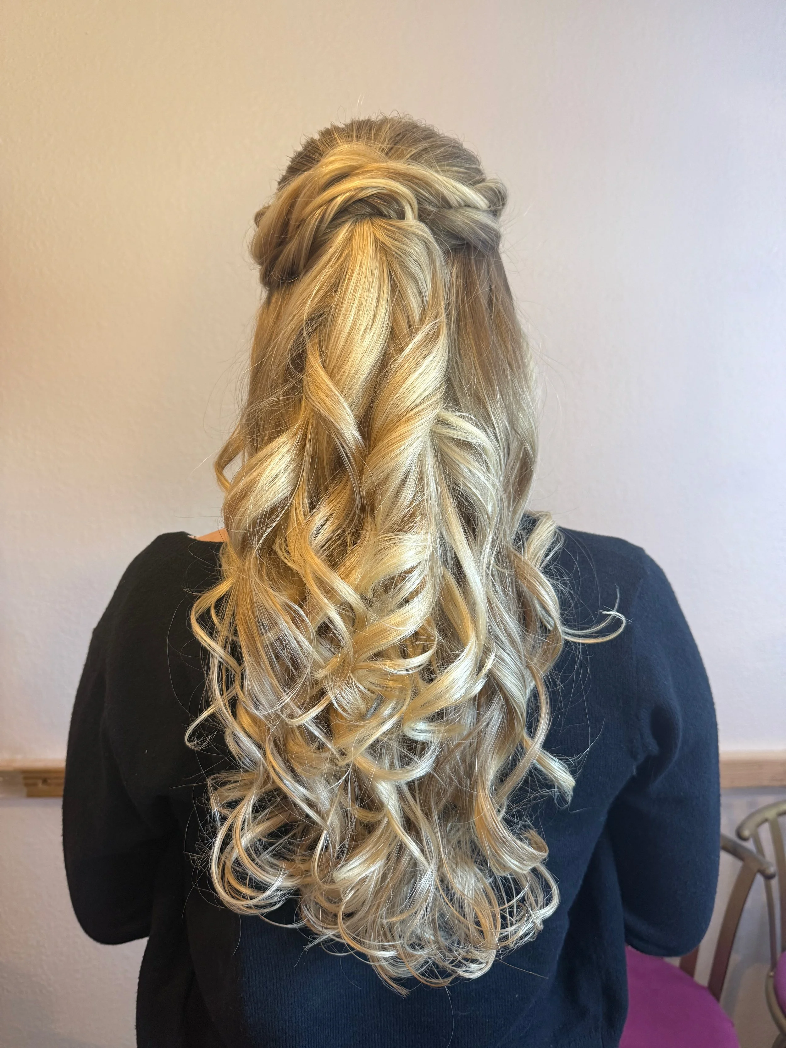 Bride with long, blonde, curly hair styled in a half-up, braided crown, wearing a black top, standing indoors against a plain wall.