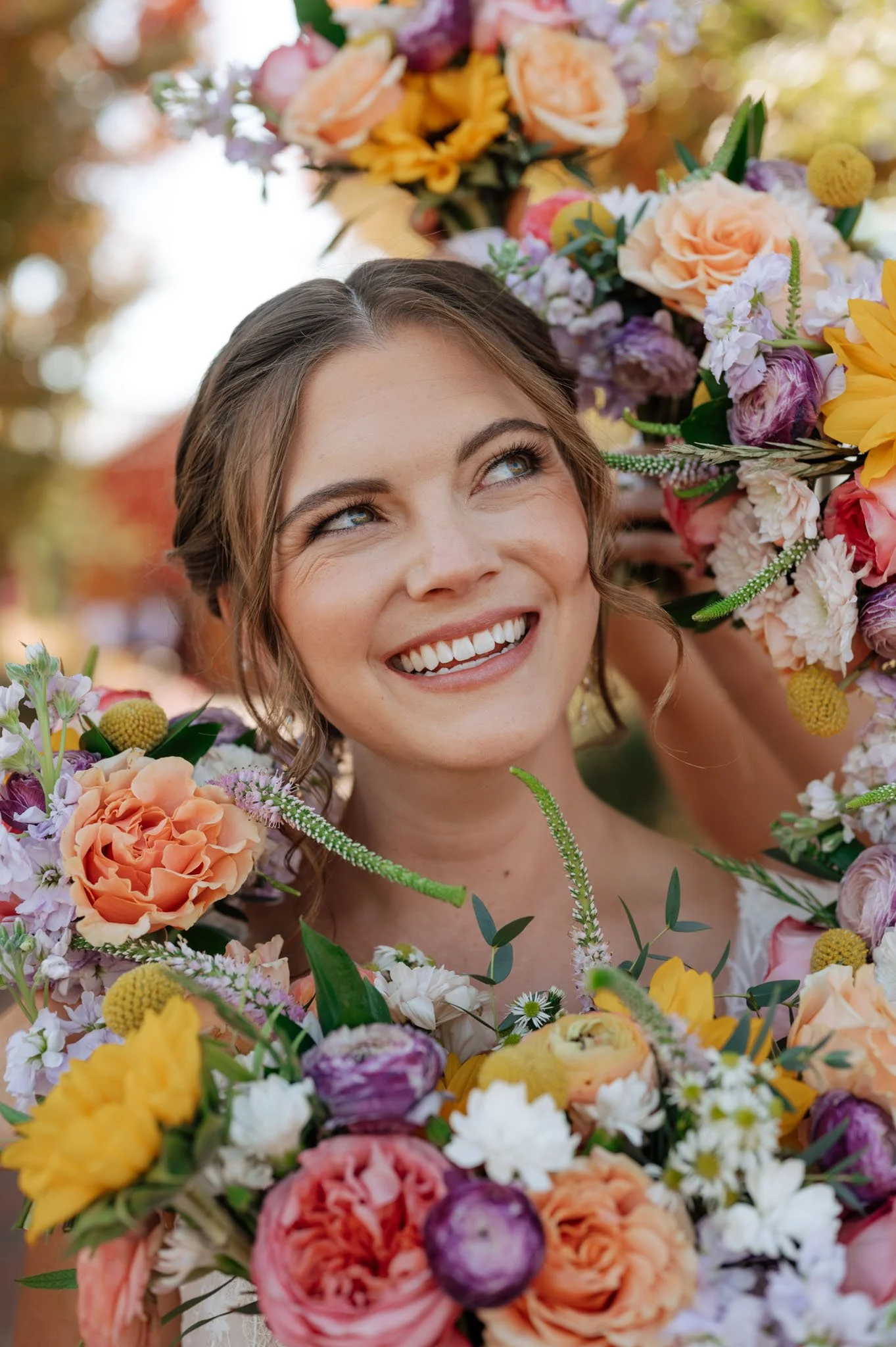 A woman with brown hair and blue eyes smiling behind a large bouquet of colorful flowers, including roses, sunflowers, and other blooms, with a blurred outdoor background.