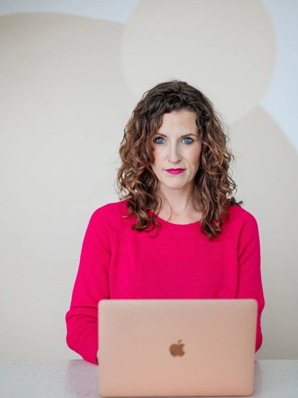 A woman with curly brown hair and blue eyes wearing a bright pink sweater, sitting at a white table with a rose gold MacBook in front of her, against a plain light-colored background with abstract shapes.