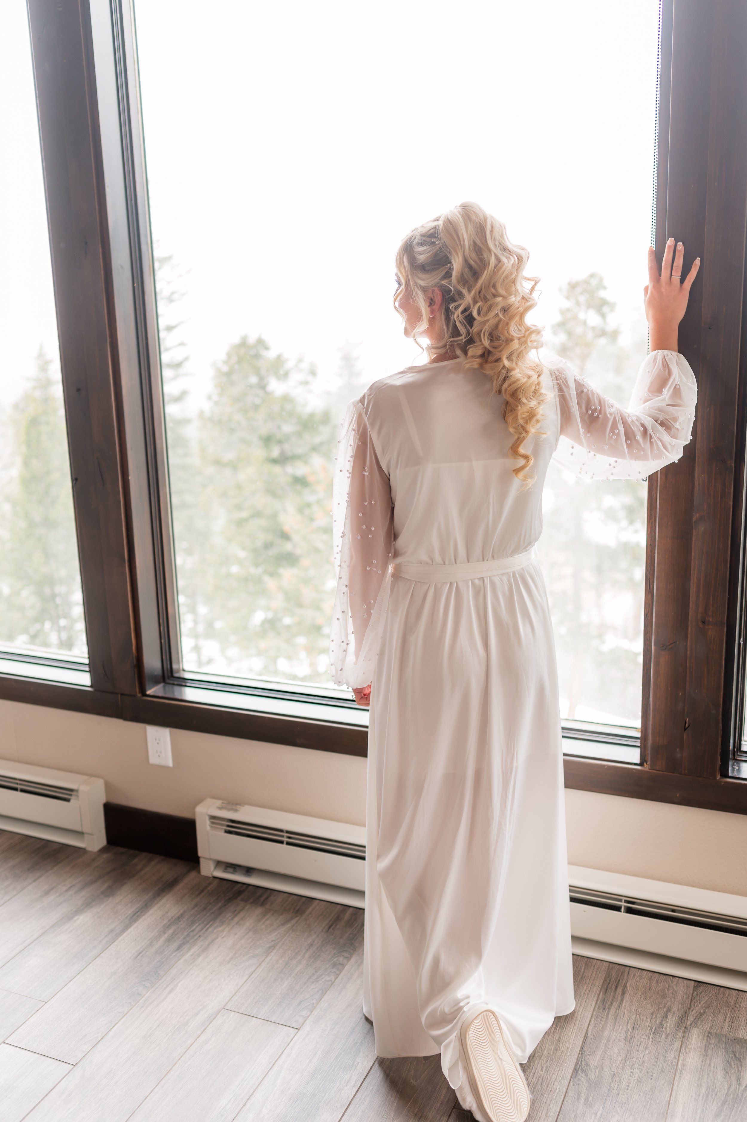 A bride with long blonde curly hair wearing hair extension,  a white dress and sneakers, standing and looking out a large window with a view of trees outside.