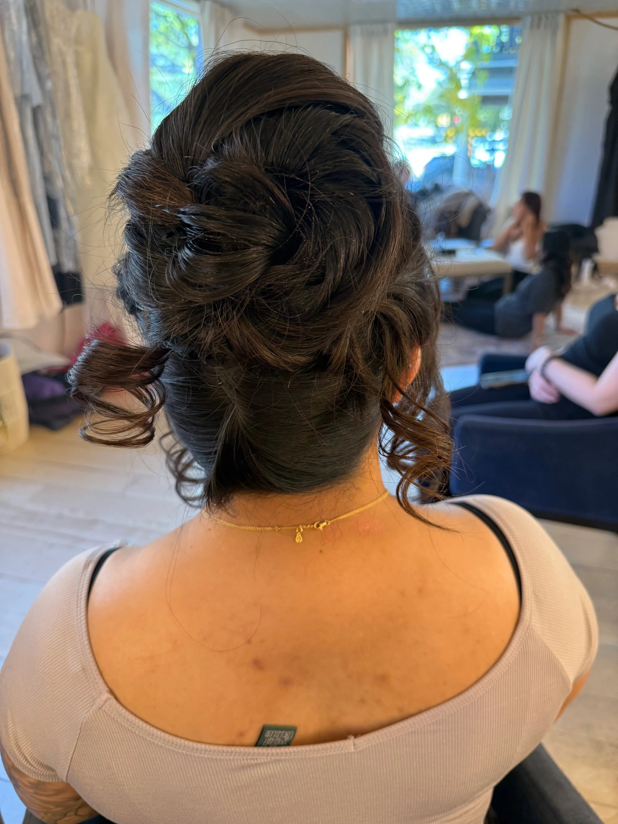 Back view of a bride with styled dark brown hair in an updo with curls, wearing a beige top with black straps and a gold necklace, in a room with large windows and people sitting in the background.