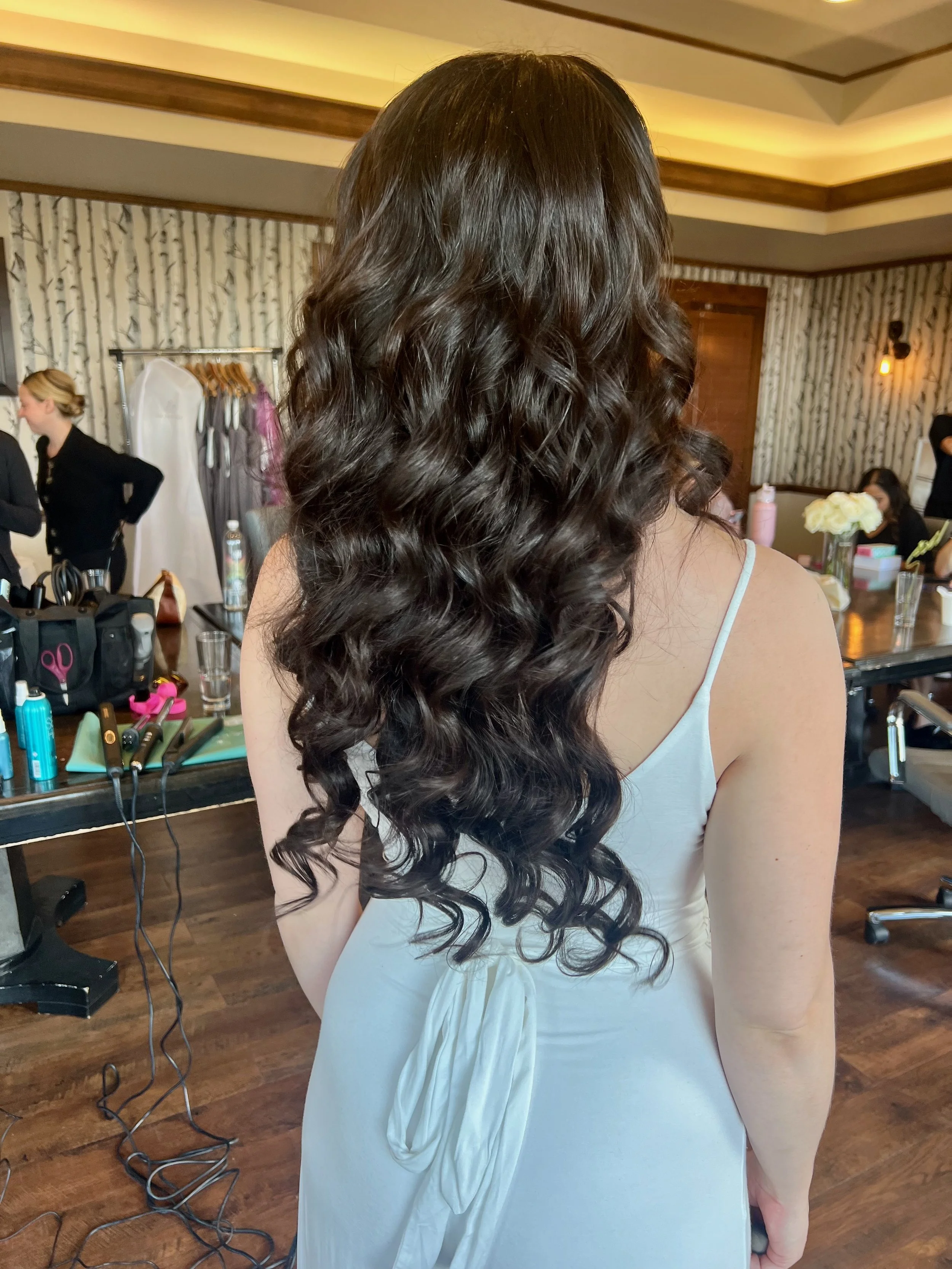 Back view of a bride with long, dark, Hollwood wave hair wearing a white sleeveless dress in a salon or dressing room with hair styling tools and products on the table.