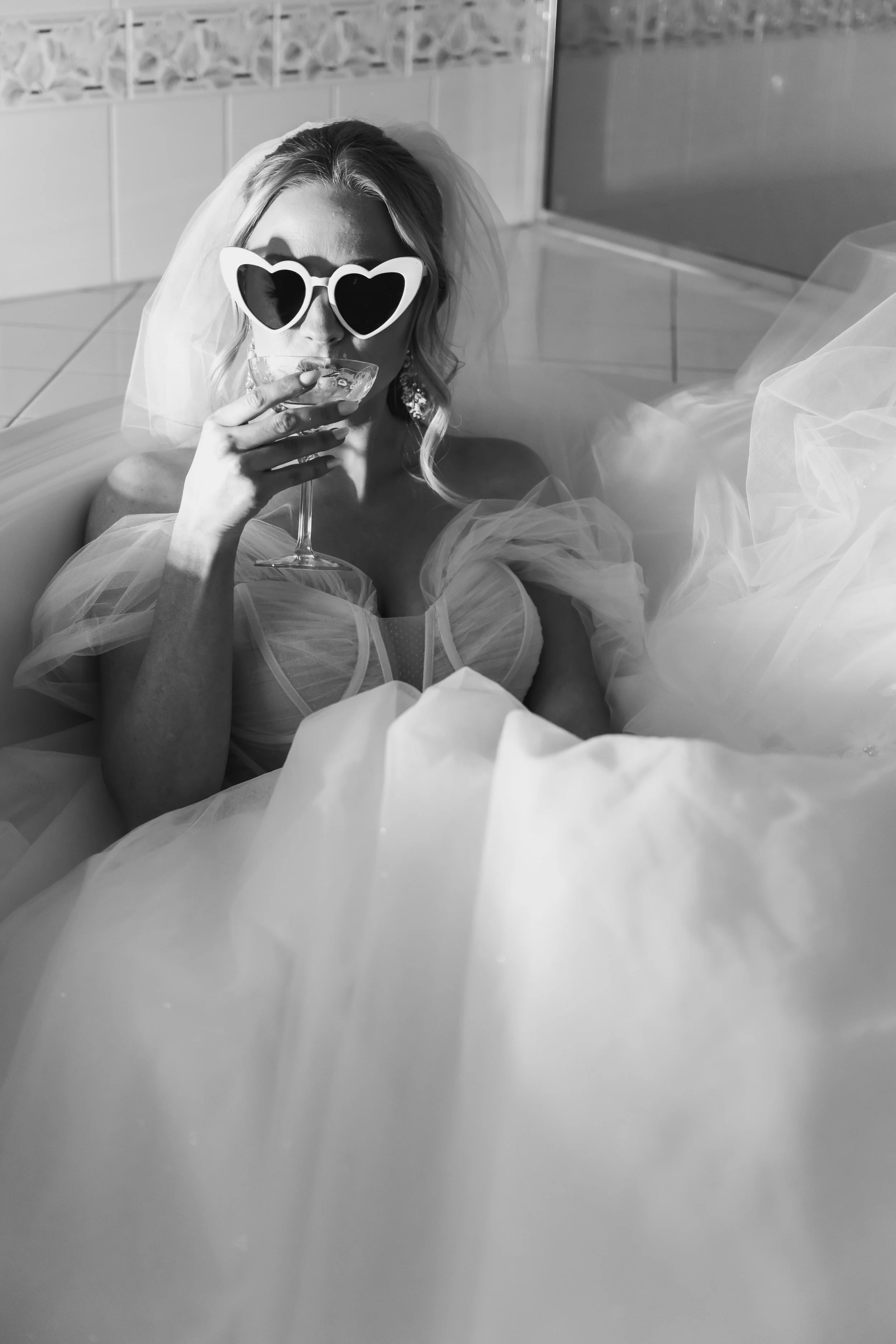 A woman wearing heart-shaped sunglasses and a bridal veil is relaxing in a bathtub, holding a glass of champagne.