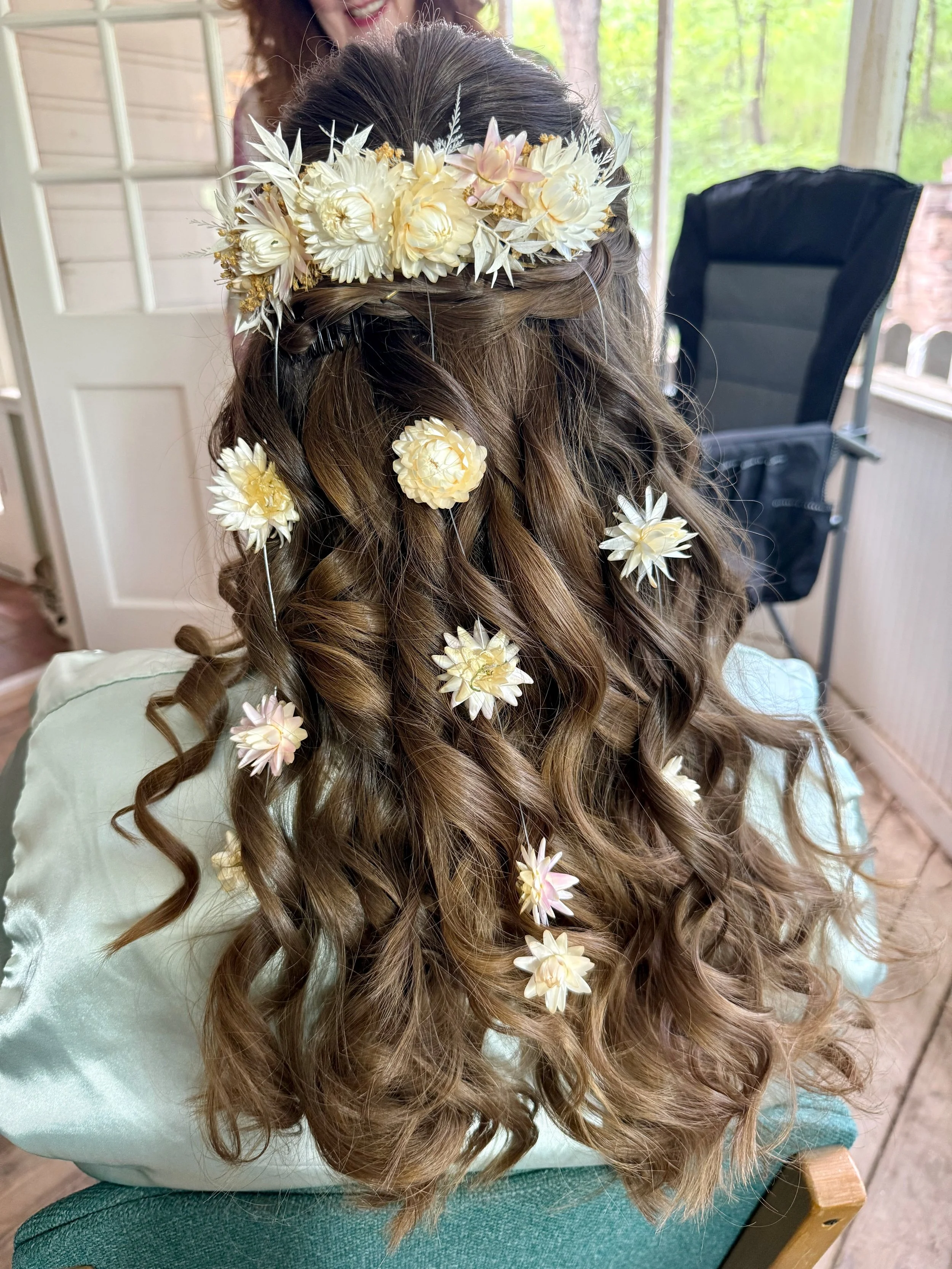 A bride with long, curly brown hair decorated with white and pale pink flowers, wearing a light-colored top, sitting on a green chair, with a person in the background on a porch or outdoor setting.