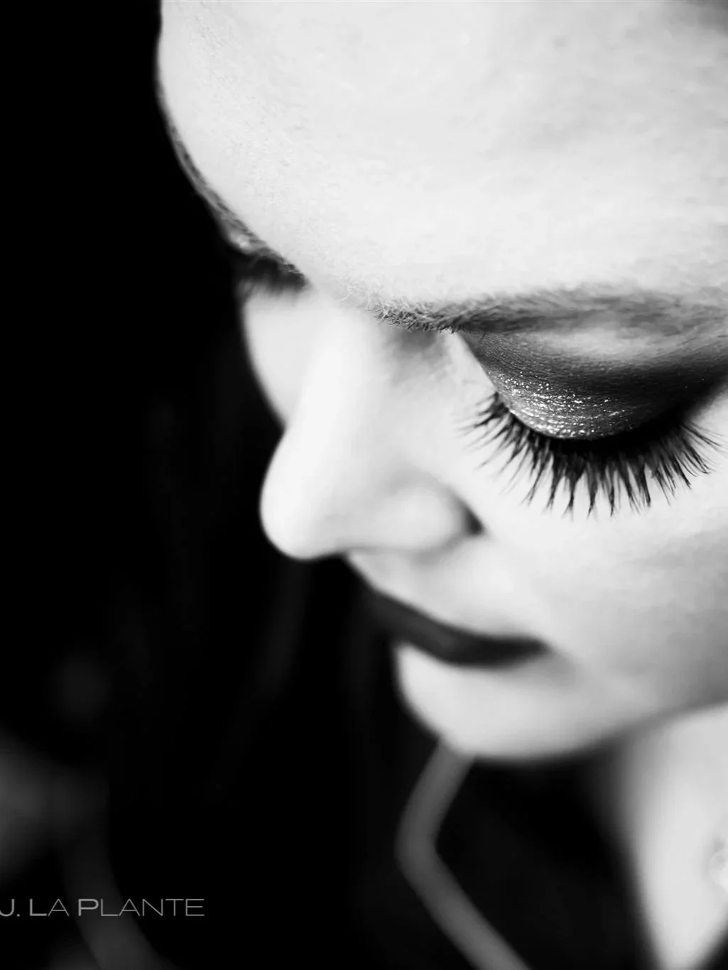 Close-up black and white photograph of a woman's face with her eyes closed, showcasing long eyelashes and shimmery eyeshadow, with a focus on her nose and eyelashes.