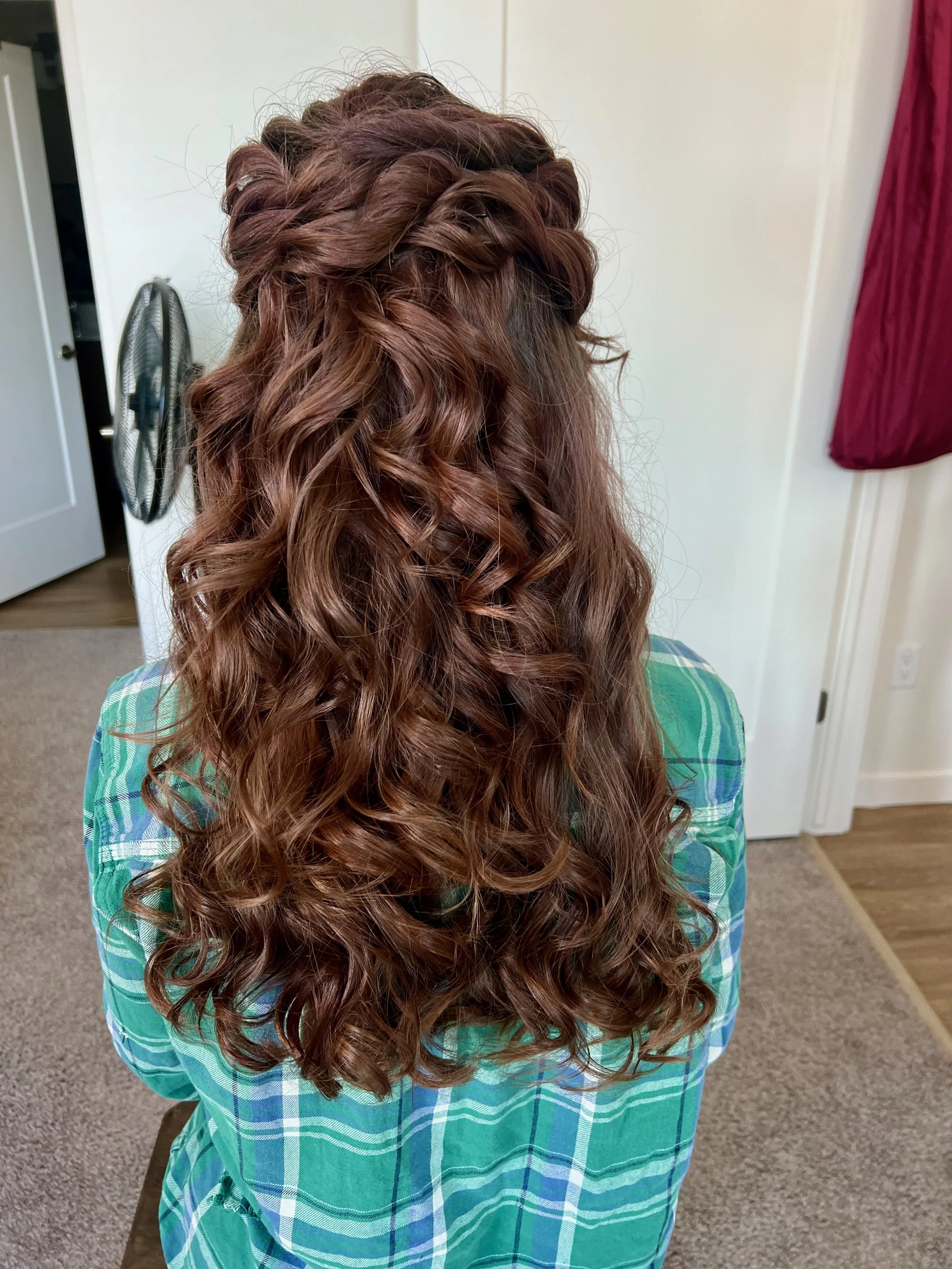 A bride with long, curly, reddish-brown hair styled with a braided crown hairstyle, wearing a green and blue plaid shirt, standing indoors with a white door, a black standing fan, and a red curtain in the background.