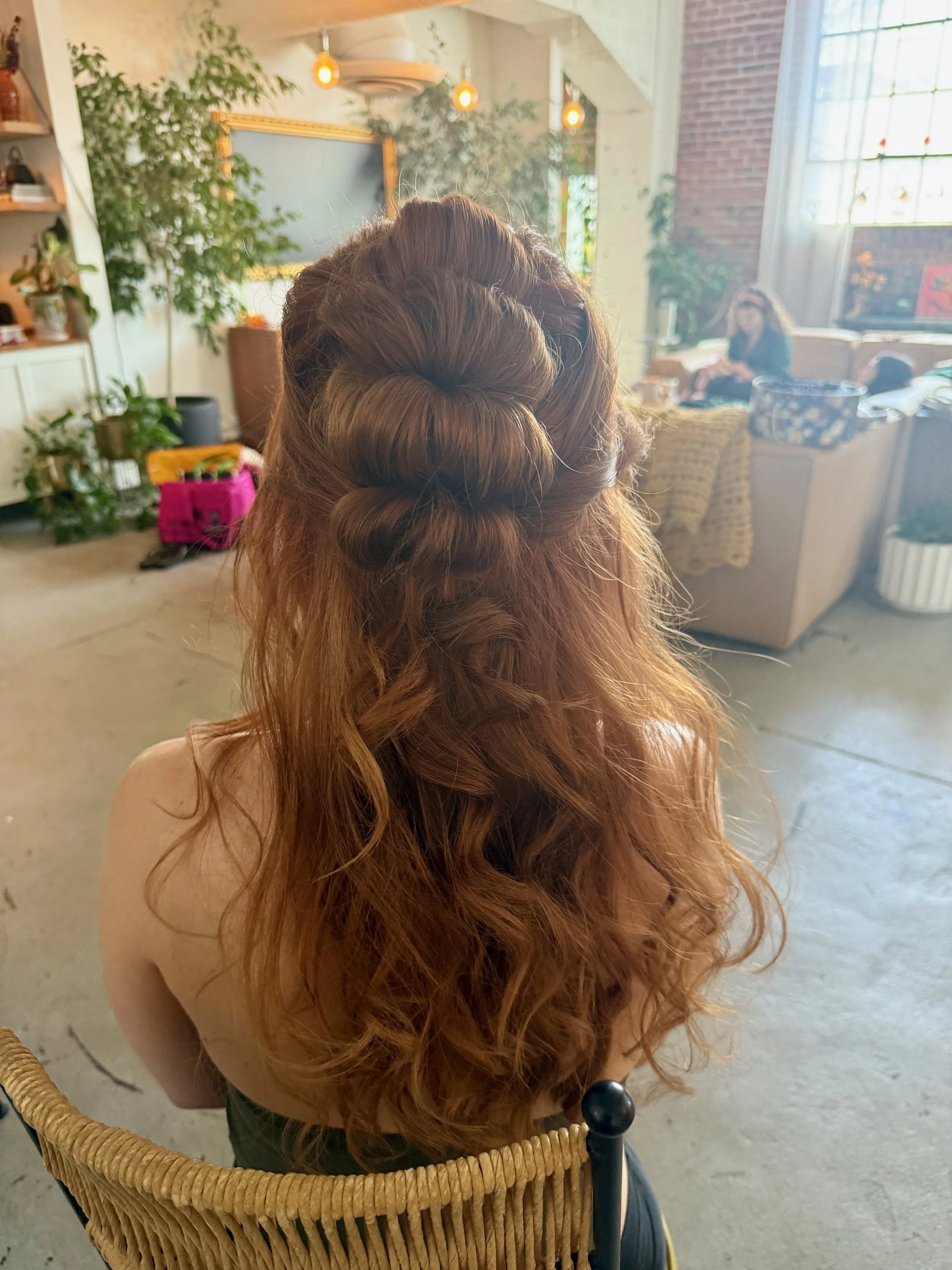 Close-up of a bride with long, red, curly hair styled in an intricate braided updo, sitting indoors in a cozy, well-lit room with large windows, plants, and furniture.