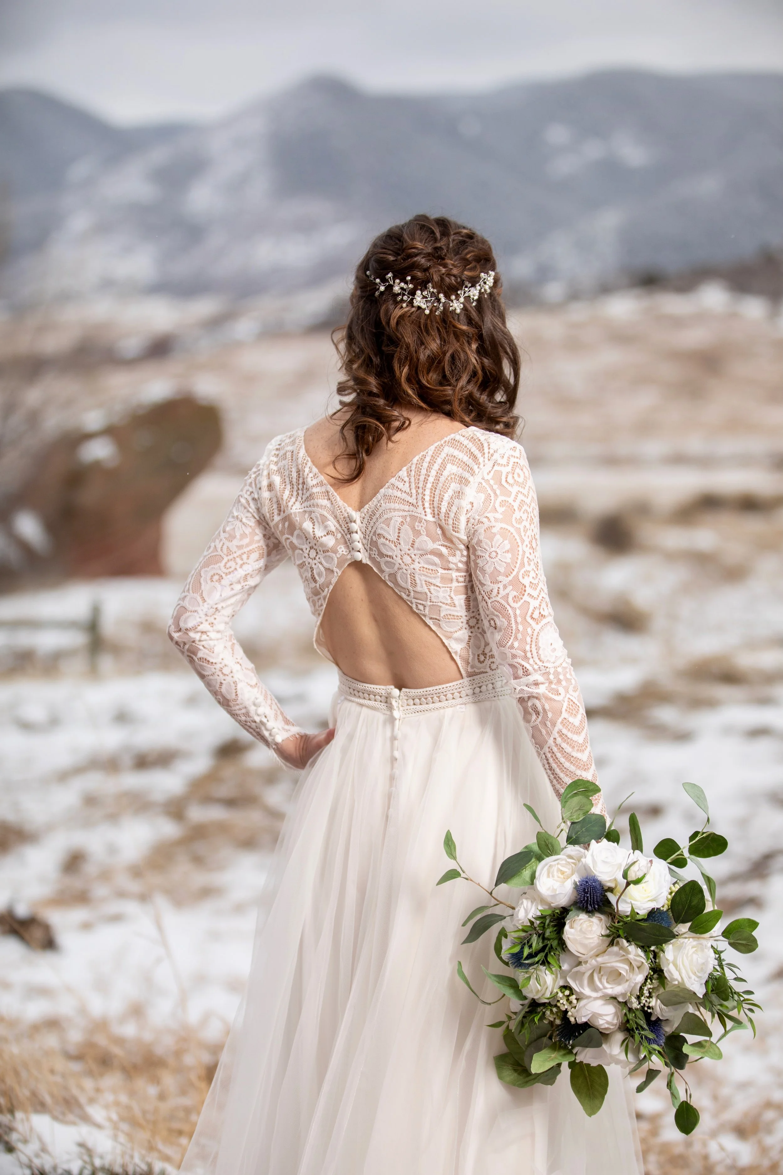 Back view of a woman in a wedding dress holding a bouquet, standing outdoors with mountains and snow in the background.