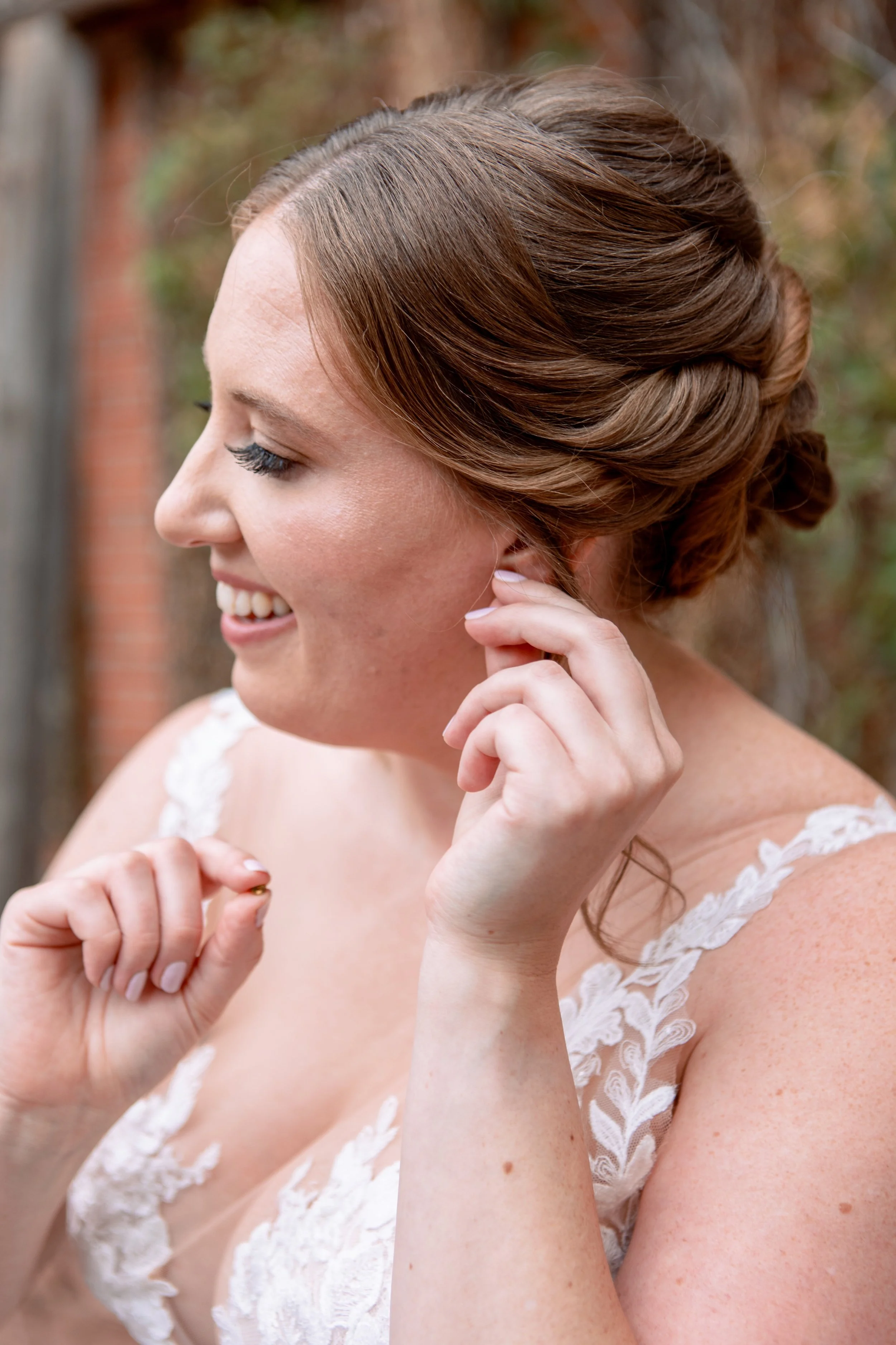 A woman with auburn hair styled in an elegant updo, wearing a white lace dress, smiling while adjusting her earring outdoors.