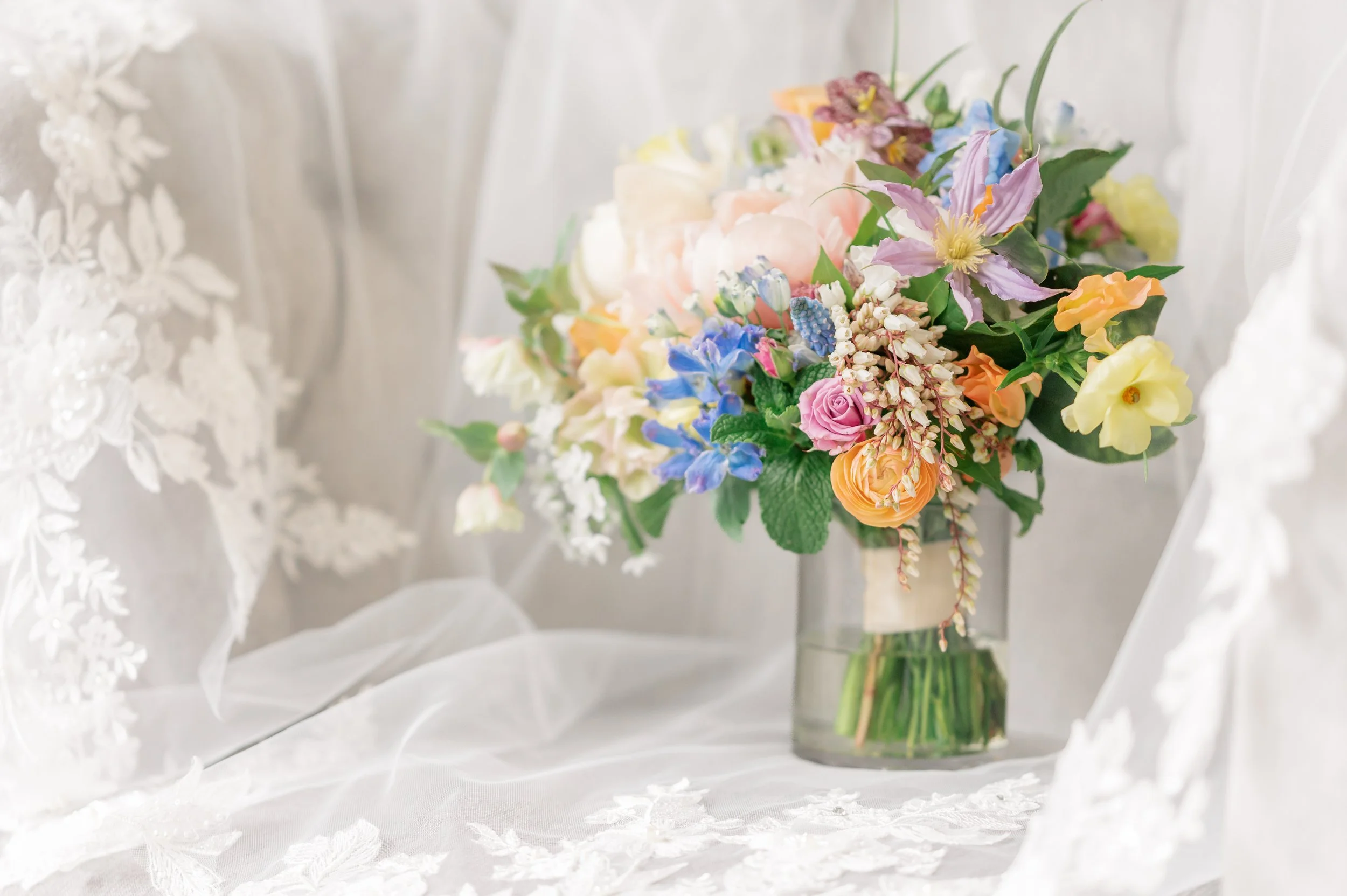 A colorful bouquet of flowers including roses, lilies, and other various flowers in a glass vase, placed on a white, embroidered fabric background.