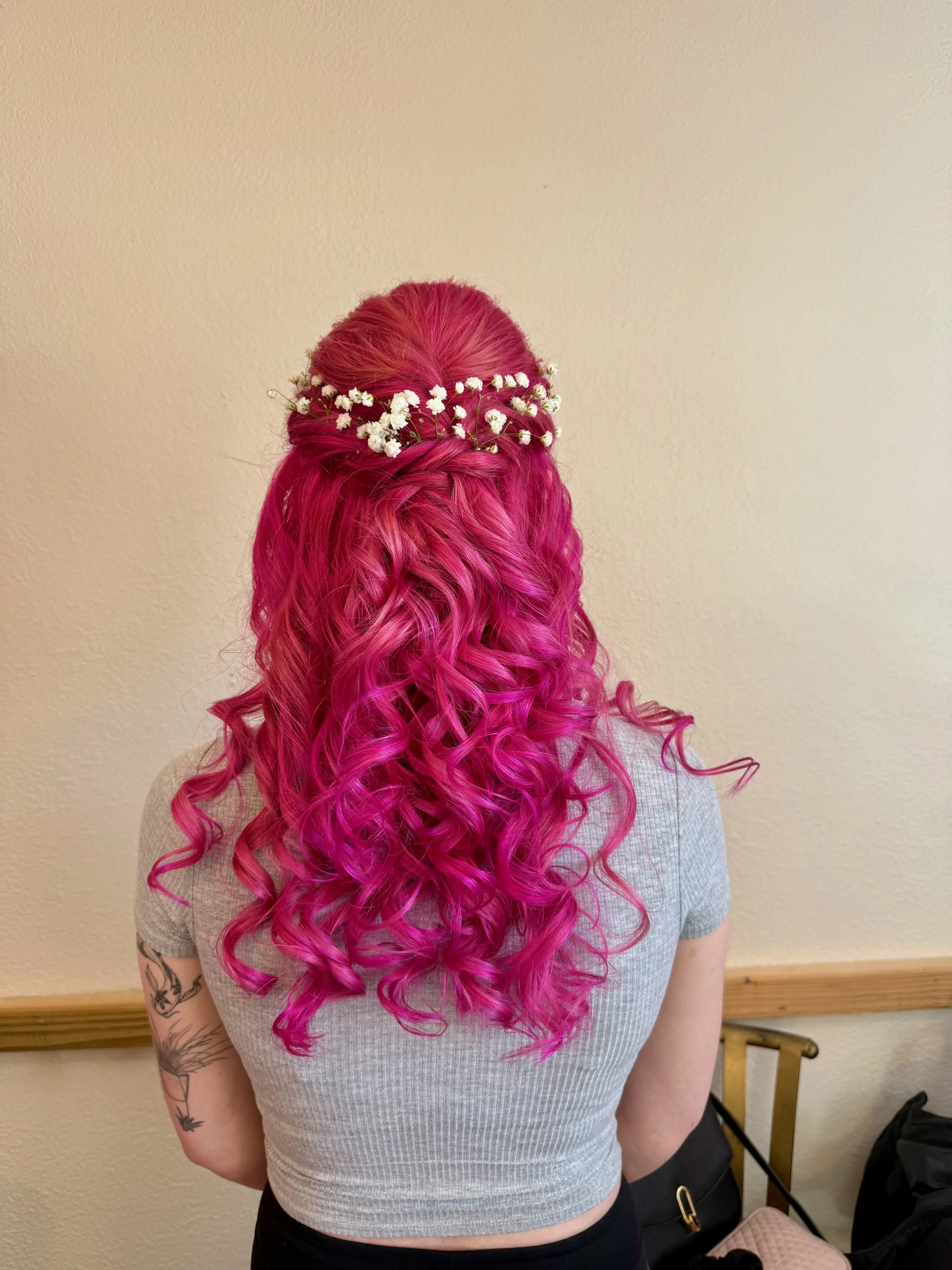 Bride with long, curly, bright pink hair wearing a floral crown made of small white flowers, sitting against a beige wall with a wooden border.