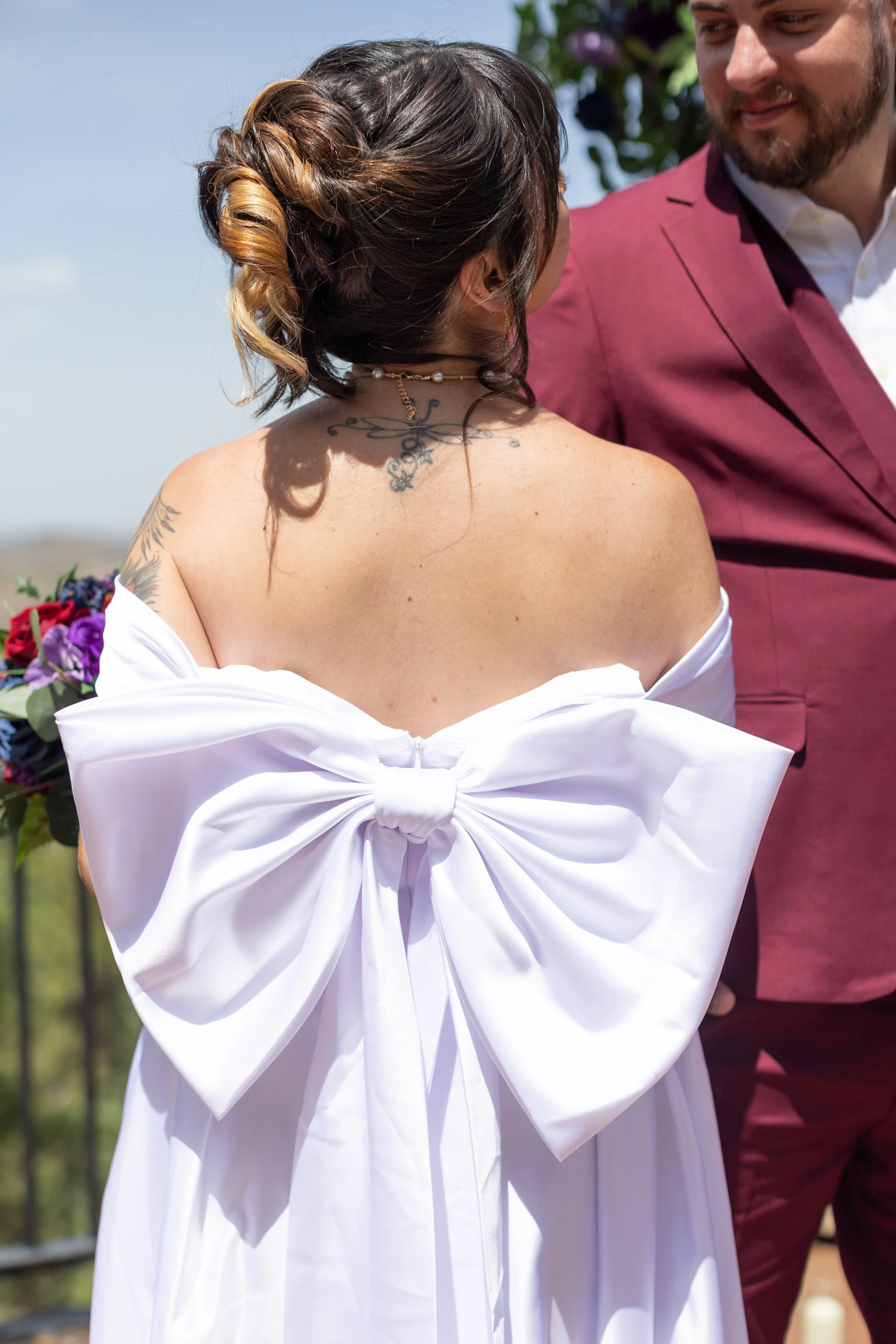 A bride in a white dress with a large bow on the back stands outdoors, facing a man in a pink suit. The woman has tattoos on her upper back and wears a pearl necklace. They appear to be at a wedding or similar celebration.