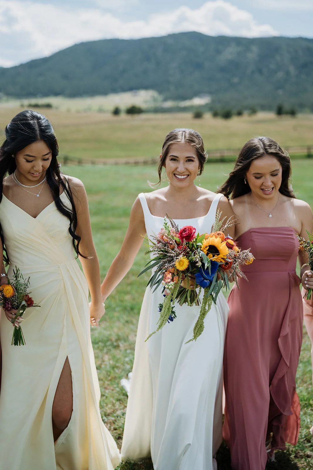 Three women in formal dresses holding hands and walking outdoors, with green grass, trees, and mountains in the background. The woman in the center is smiling and holding a colorful bouquet of flowers.