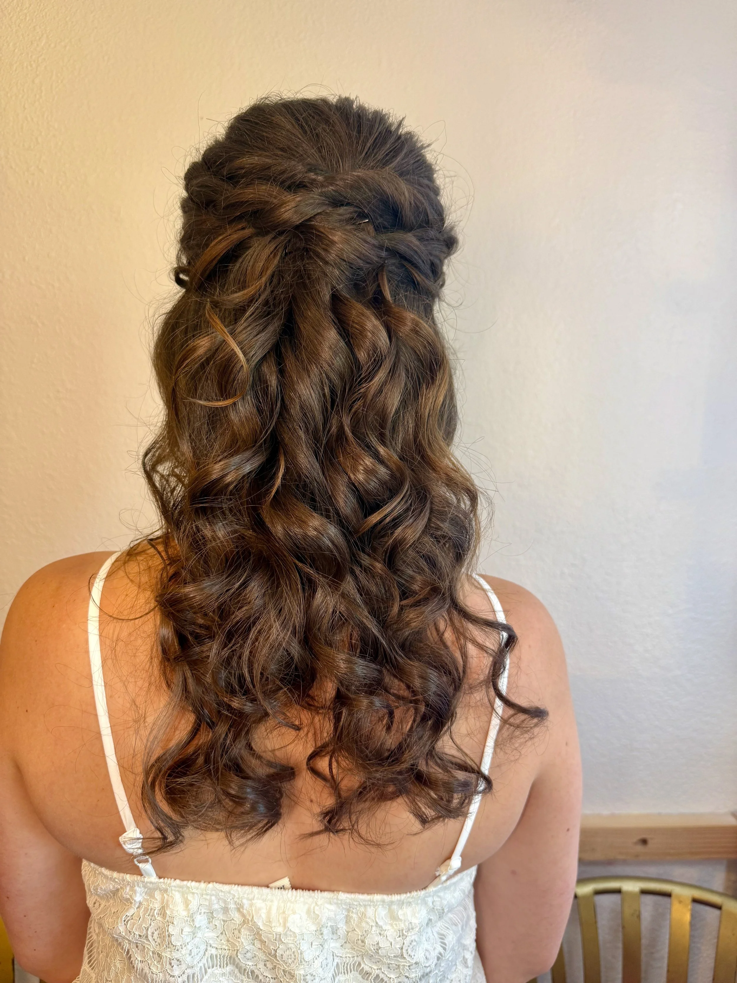 Back view of a bride with long, curly brown hair styled in loose waves, wearing a white lace dress with spaghetti straps, standing indoors against a plain light-colored wall.