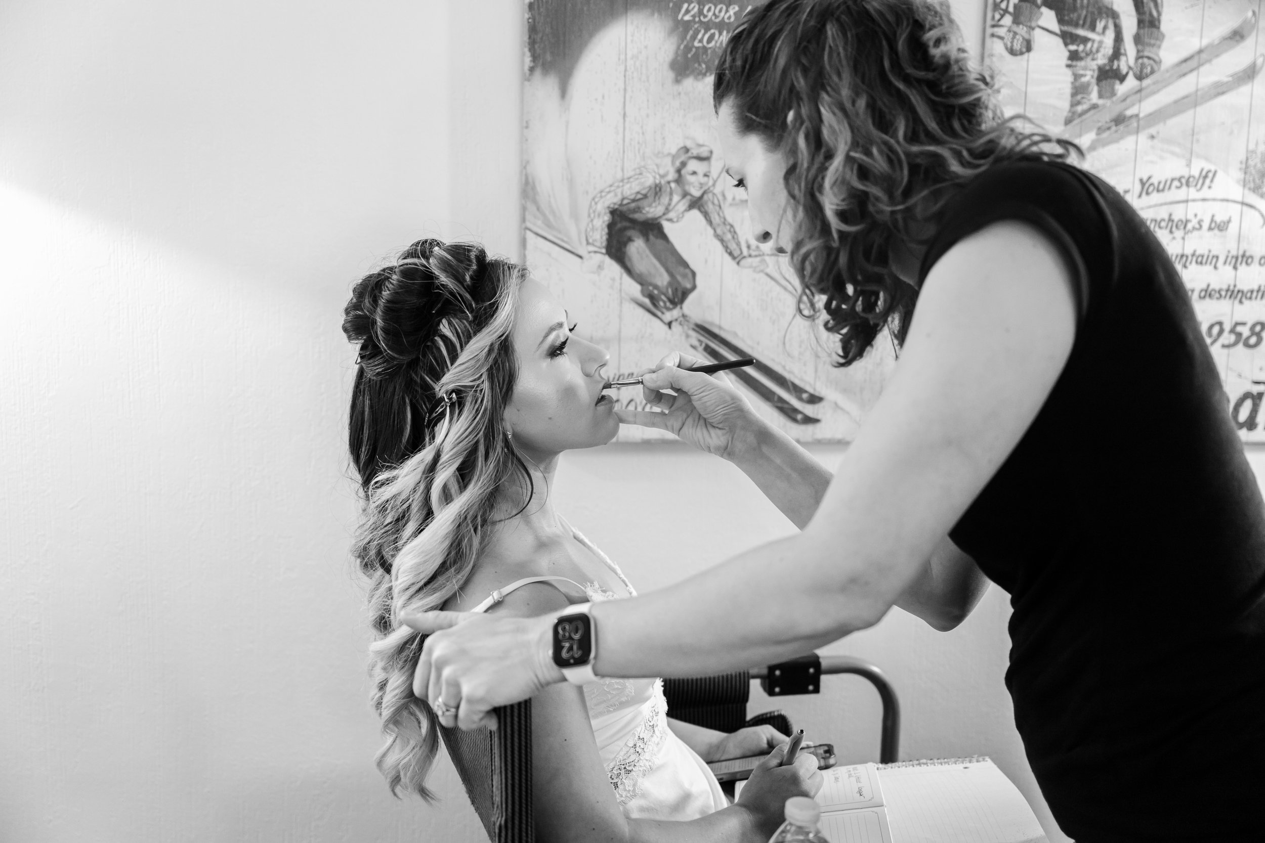 A makeup artist applies lipstick to a woman sitting in a chair, holding a mirror and a makeup brush. The woman has styled hair and makeup, and a notebook and water bottle are on the table in front of her. There is a painting of a skateboarder on the wall behind them.
