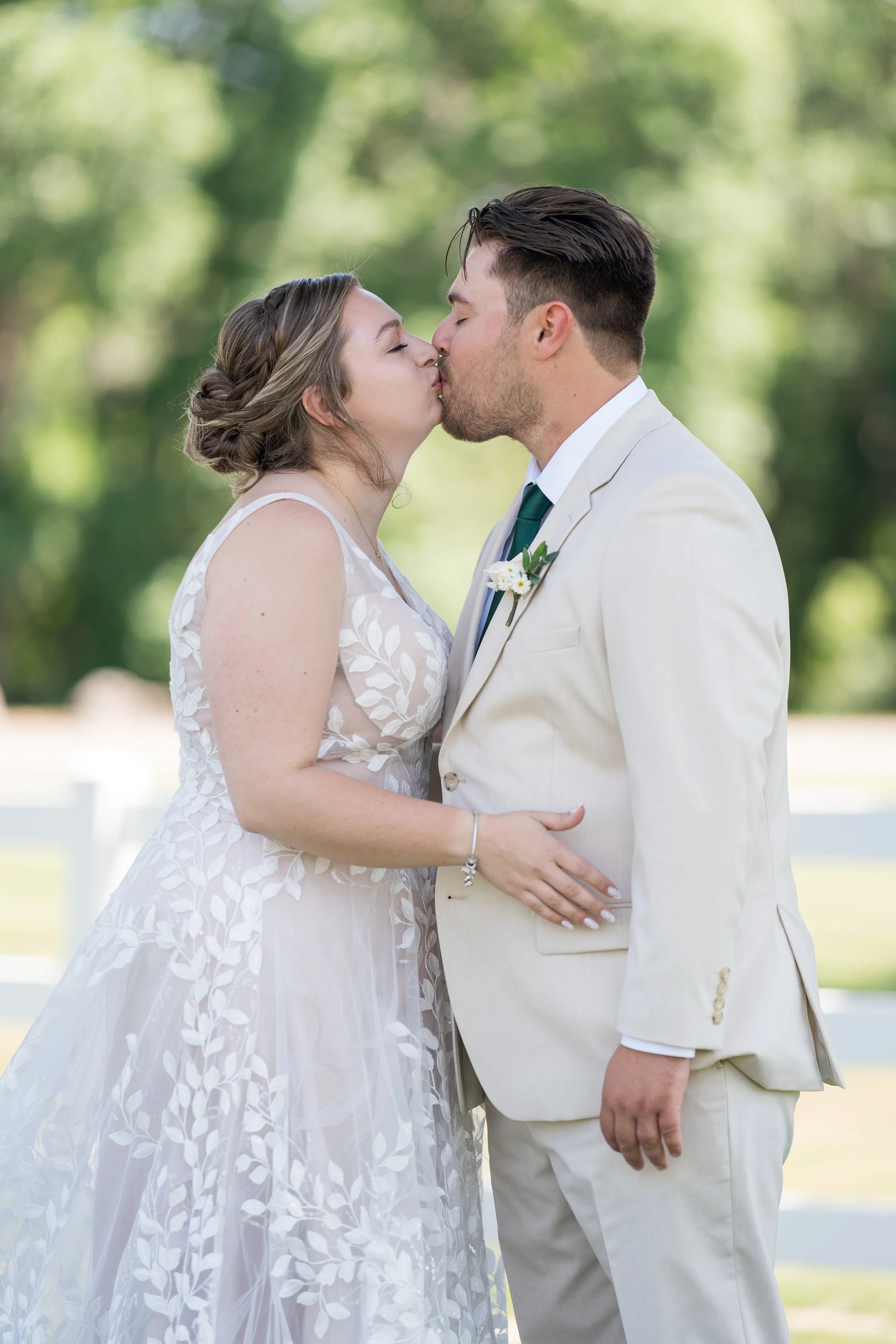A bride and groom sharing a kiss outdoors during their wedding, with greenery in the background.
