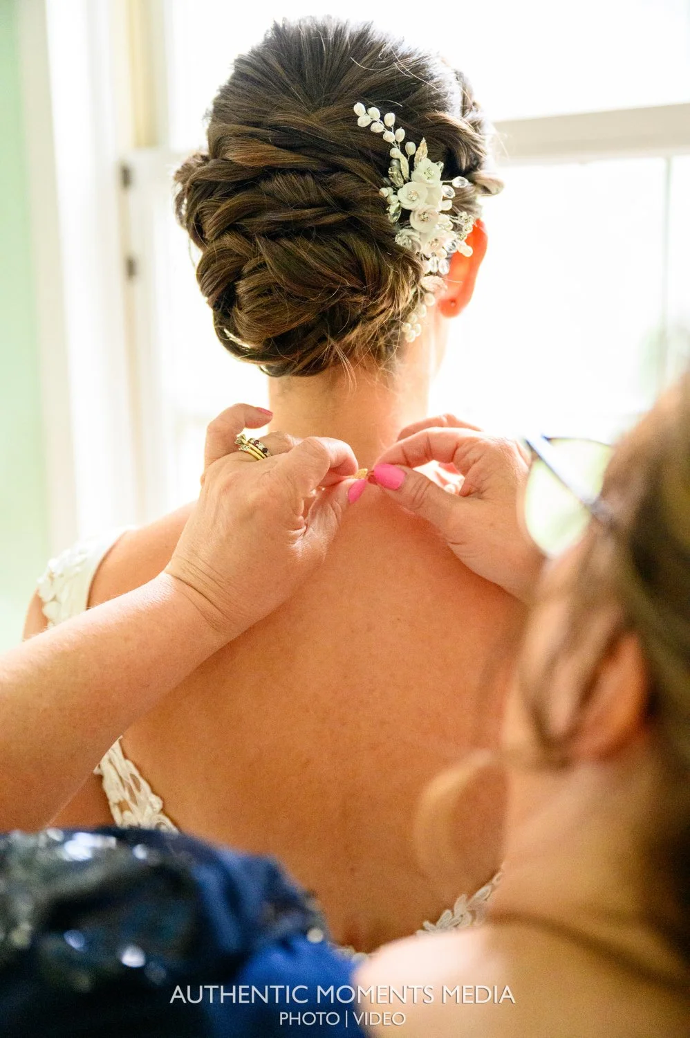 A bride getting ready with her hair styled in an elegant updo decorated with white floral accessories, while someone adjusts her strapless dress.