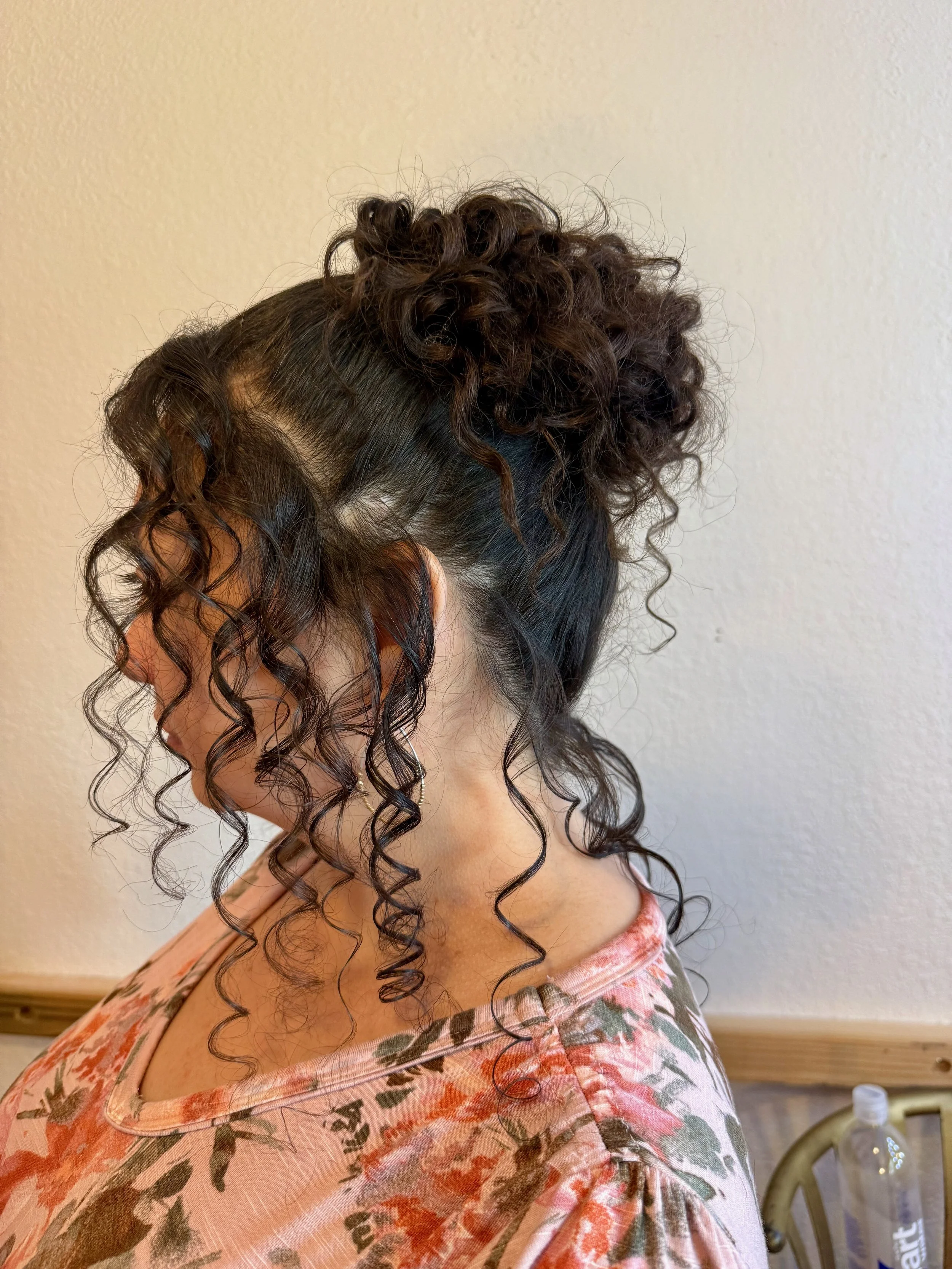 Close-up of a bride's curly hair styled in an updo with tight curls and some strands hanging loose, against a plain wall background.