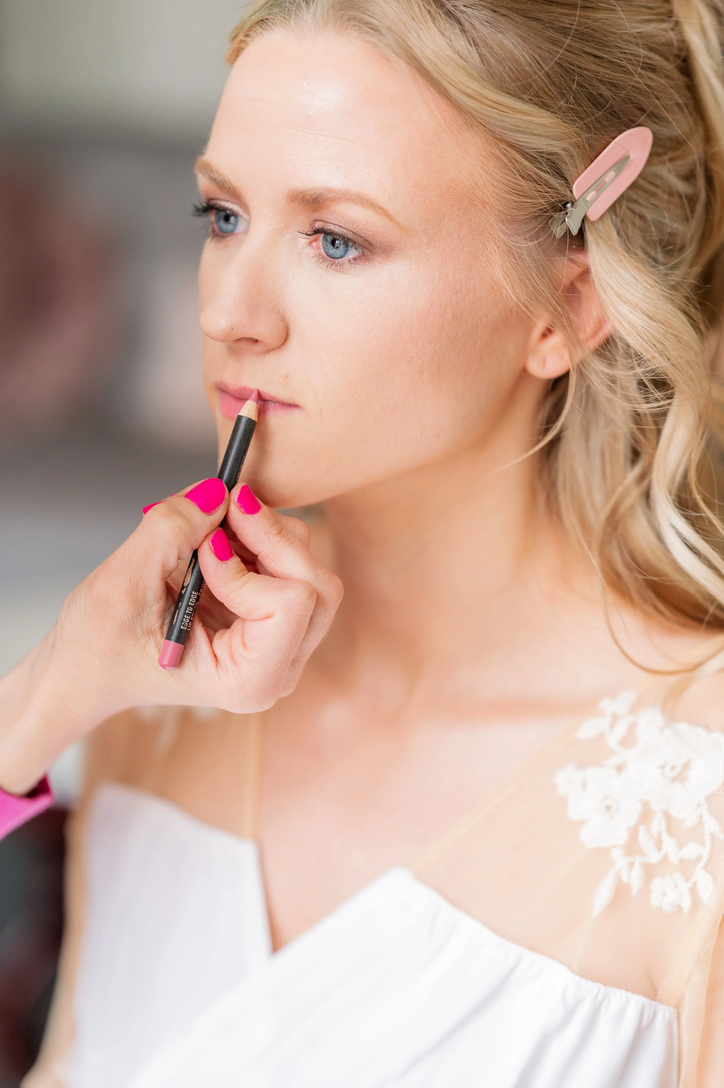 A woman with blonde hair clipped back with a pink hair clip is applying lipstick while wearing a white dress with lace details on the shoulder.