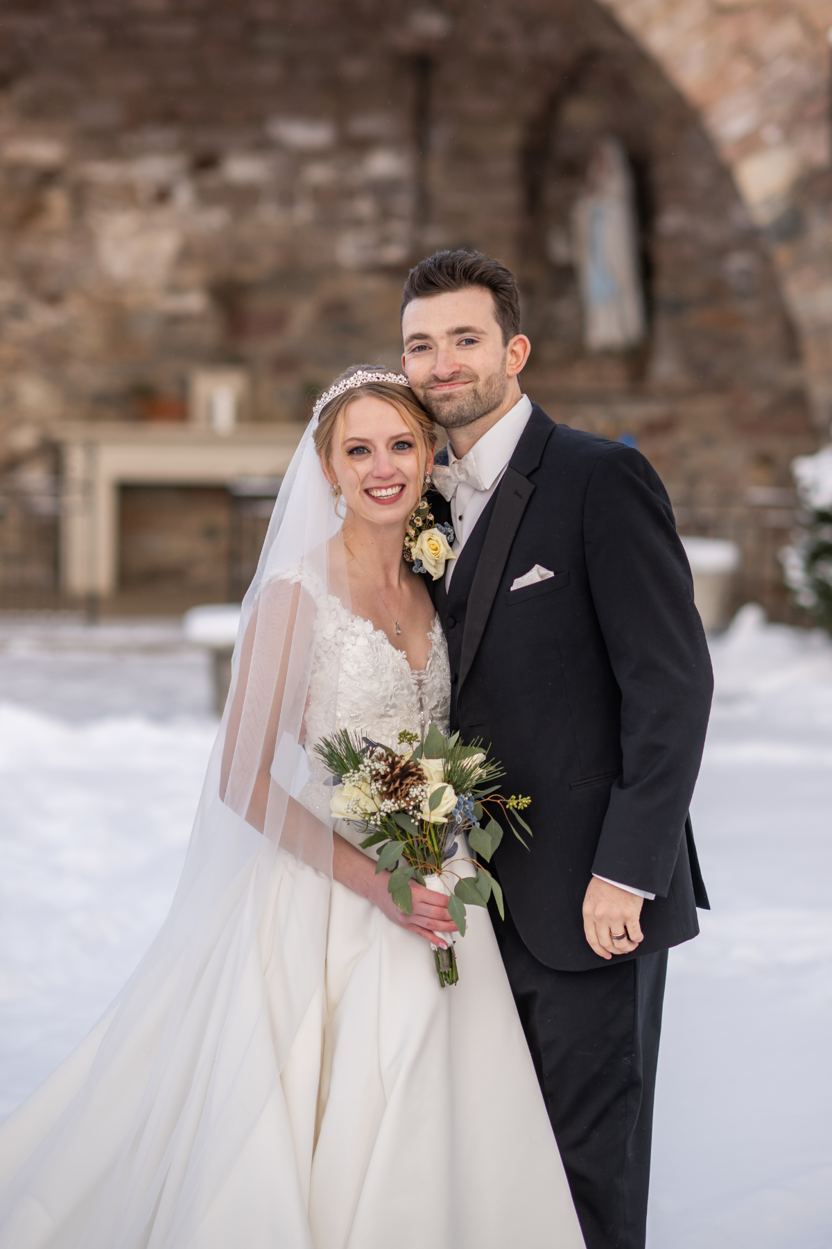 A bride and groom smiling outdoors in a snowy setting, with the bride in a white wedding dress and veil holding a bouquet, and the groom in a black tuxedo with a white shirt and bow tie, standing close together.