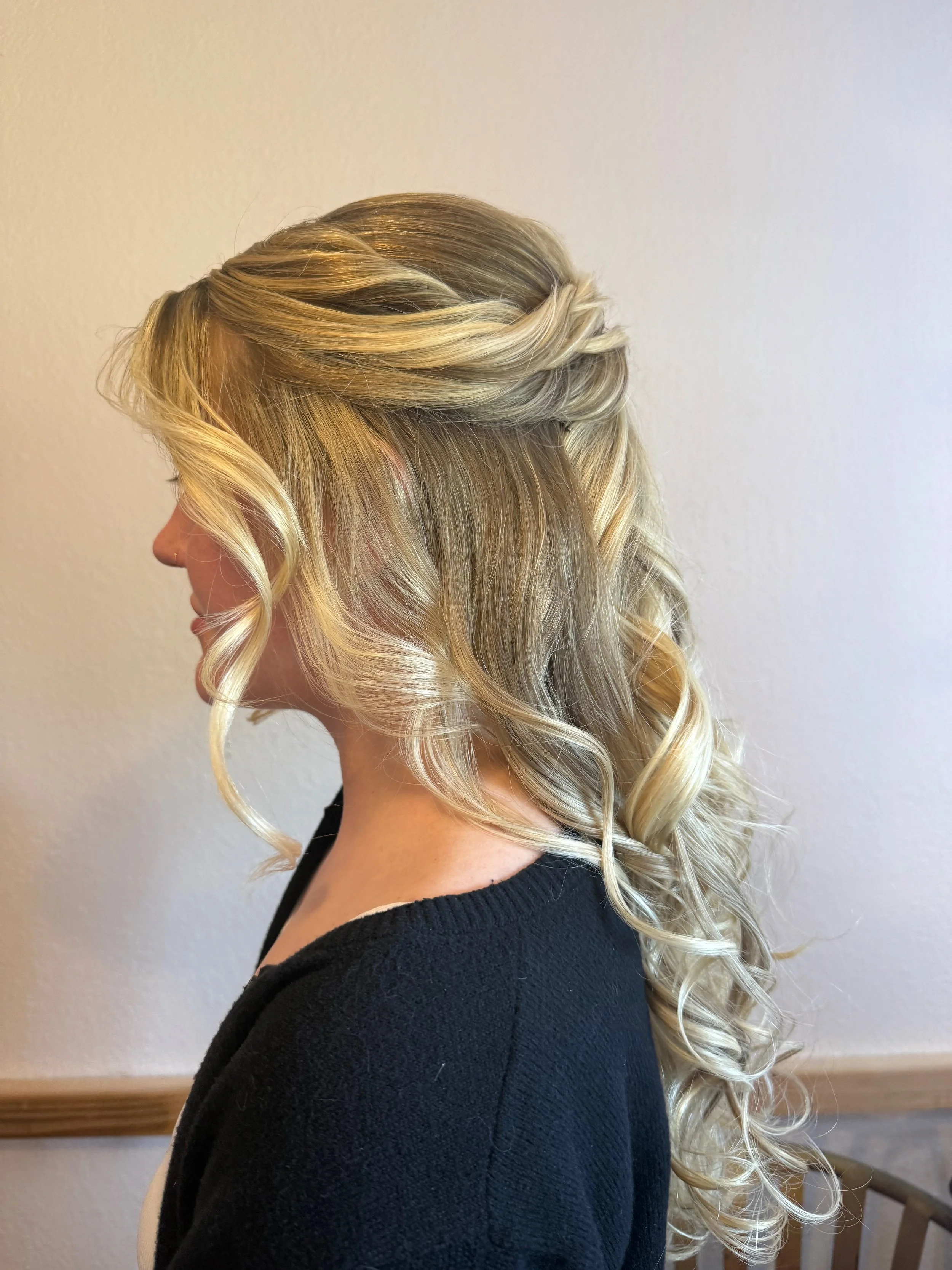 Side view of a bride with blonde, curled hair wearing a black sweater, standing next to a light-colored wall.