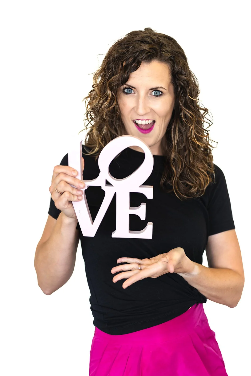 A woman with curly brown hair and blue eyes, wearing a black top and pink pants, holding a pink and white 'LOVE' sign and smiling with her mouth open.