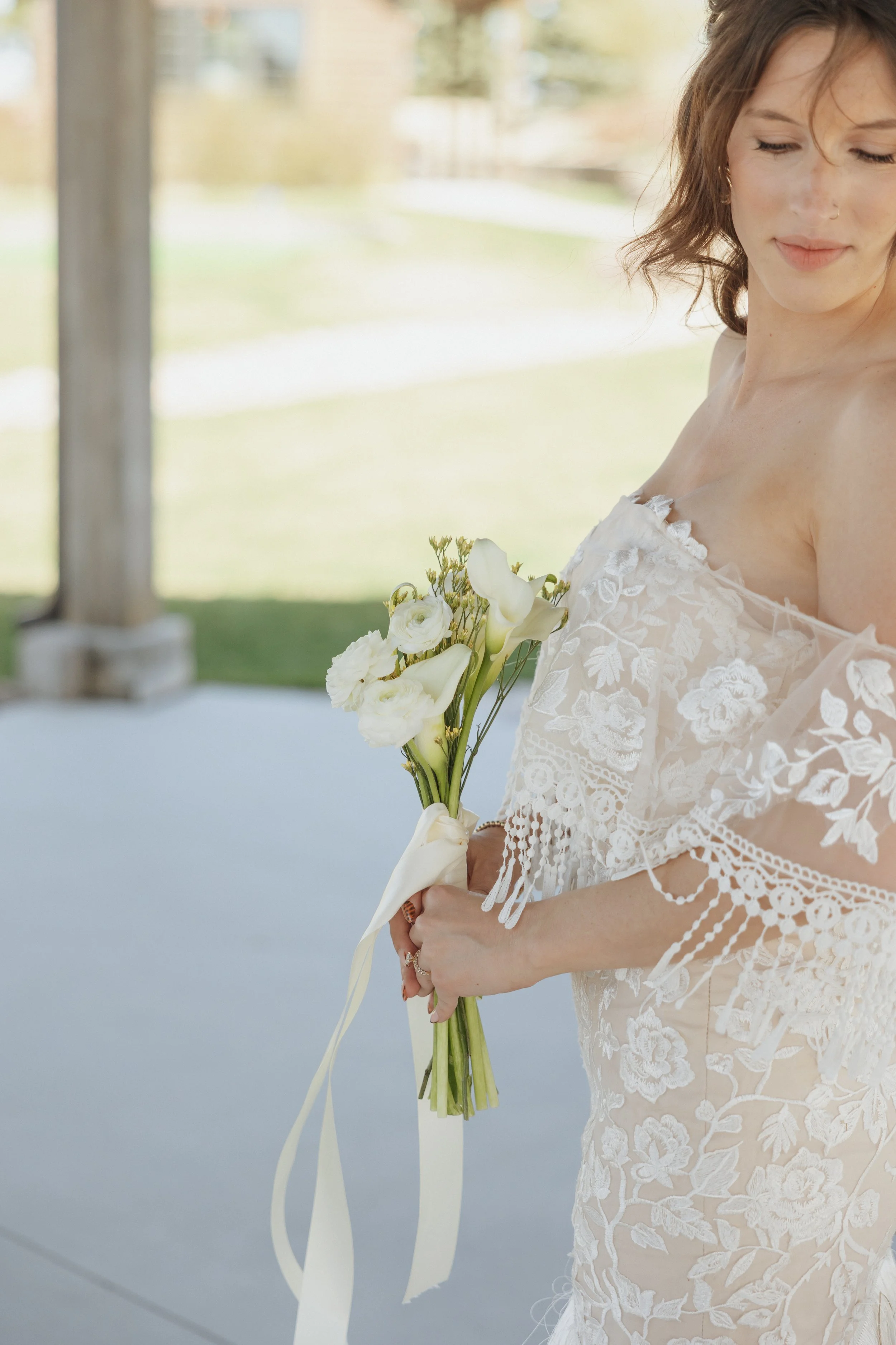 A woman in a wedding dress holding a bouquet of white flowers, standing outdoors in a bright setting.