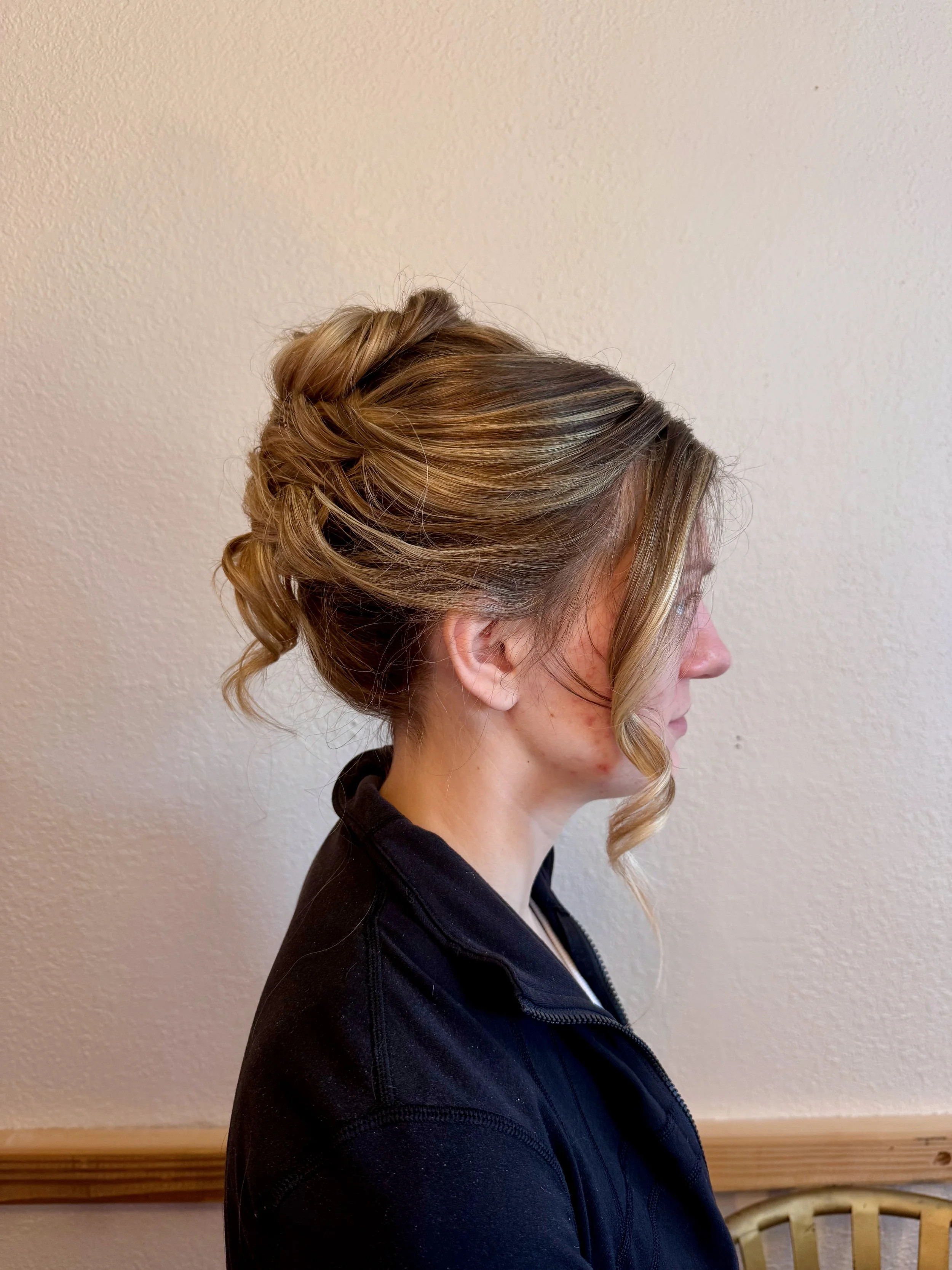 Bride with blonde and light brown hair styled in an elegant updo, wearing a black jacket, standing against a beige wall with a wooden ledge and chair in the background.